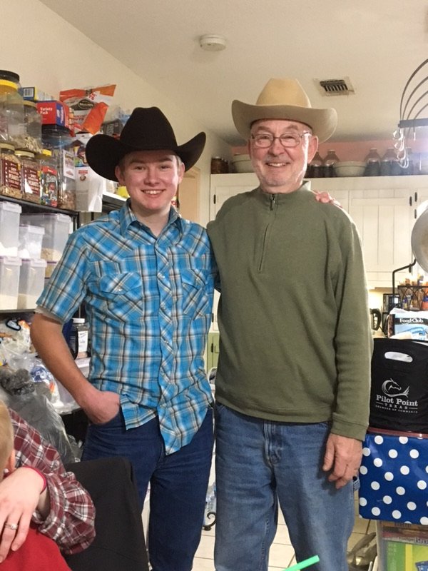 Two males, one younger and one older, wearing cowboy hats and smiling indoors. The younger wears a blue plaid shirt, and the older wears a green pullover and glasses. They appear to be in a kitchen or pantry area.