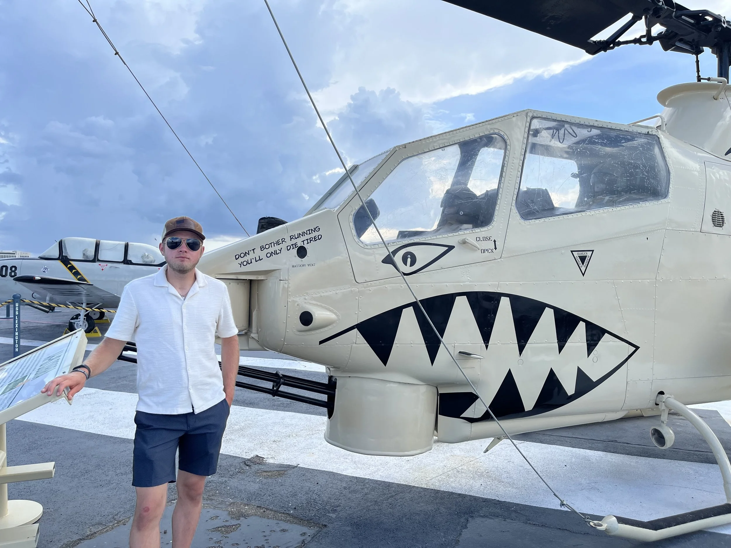 A man wearing sunglasses, a white shirt, and shorts standing next to a military helicopter with shark mouth nose art, on an airfield under a partly cloudy sky.