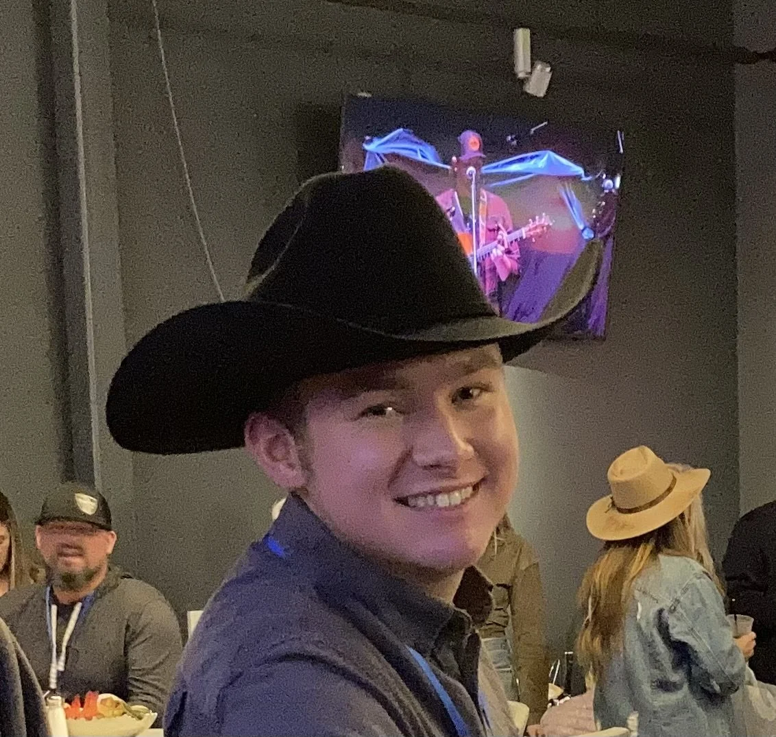 A young person smiling while wearing a large black cowboy hat at an indoor event with other people, some wearing hats, and a television screen showing a person playing guitar in the background.