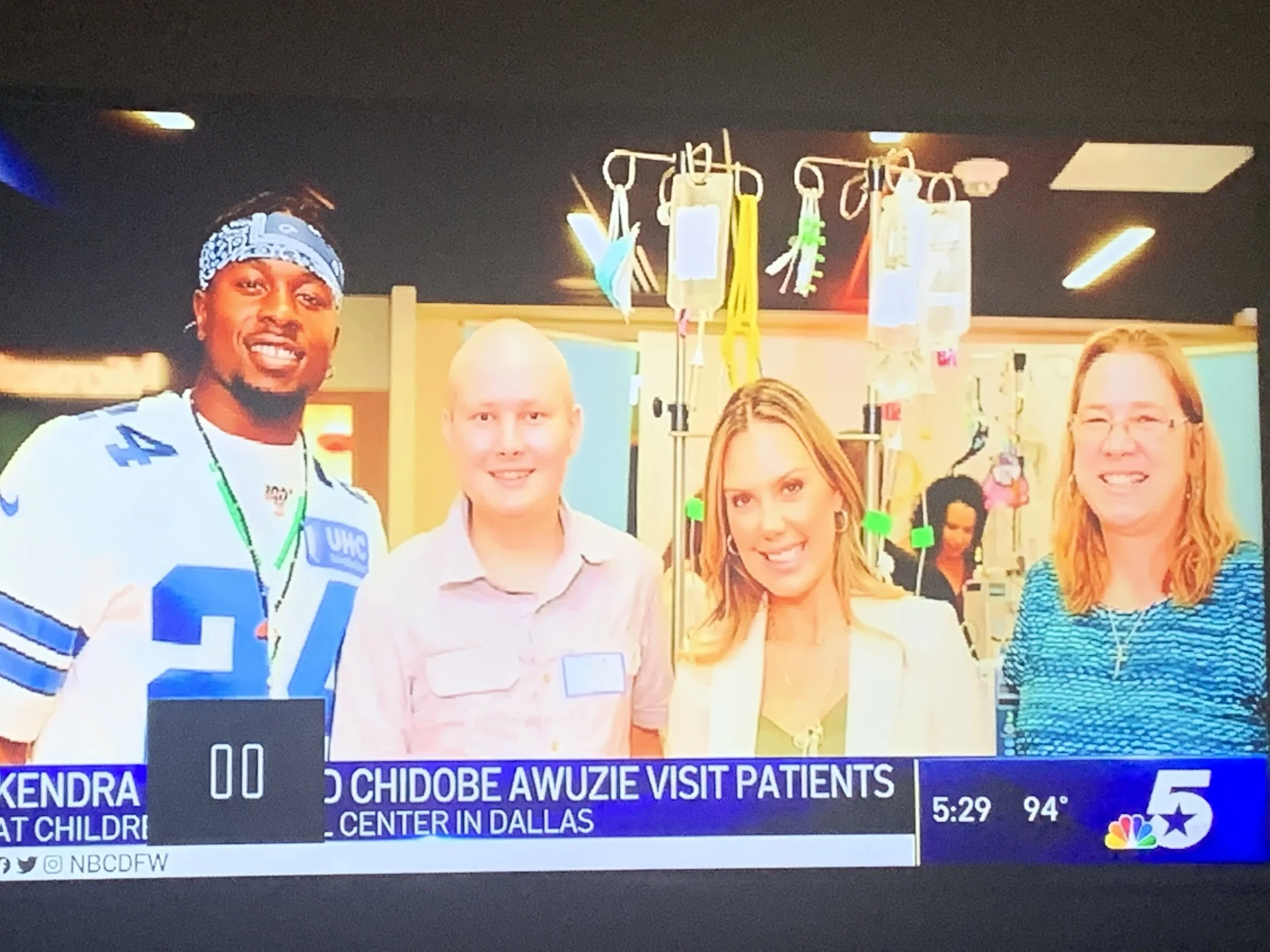 Group of four people smiling in a hospital or clinic setting with medical equipment and supplies in the background. Text overlay mentions a visit at Texas Children's Hospital in Dallas.