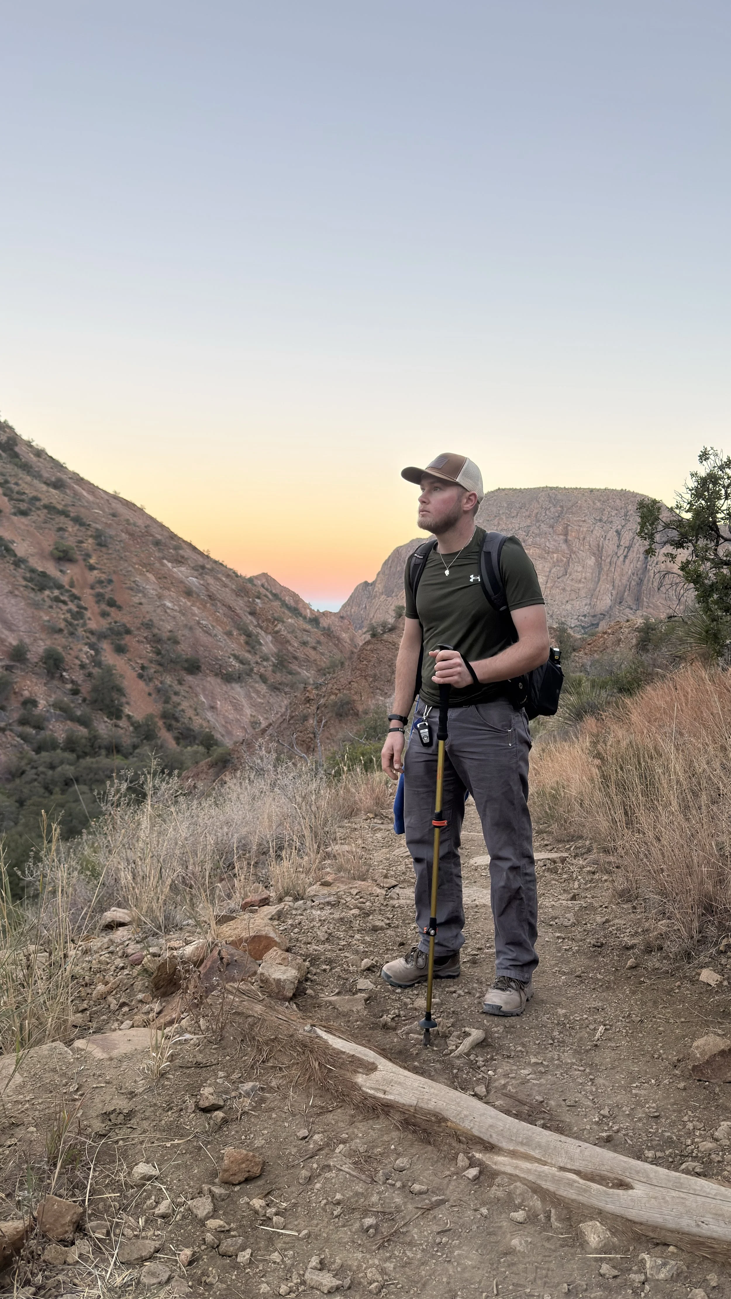 A man standing on a hiking trail in a desert canyon at sunset. He is wearing a cap, backpack, and outdoor gear, holding a hiking pole.