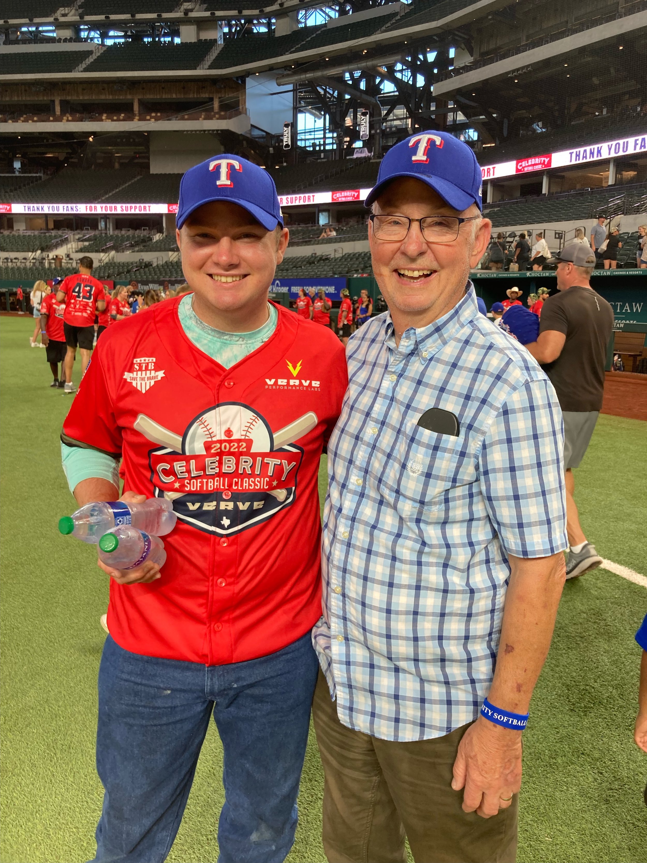 Two men smiling at a sports event inside a stadium, both wearing blue caps with a red and white logo, and the younger man in a red sports jersey with logos and text. Several people are in the background on a baseball field.