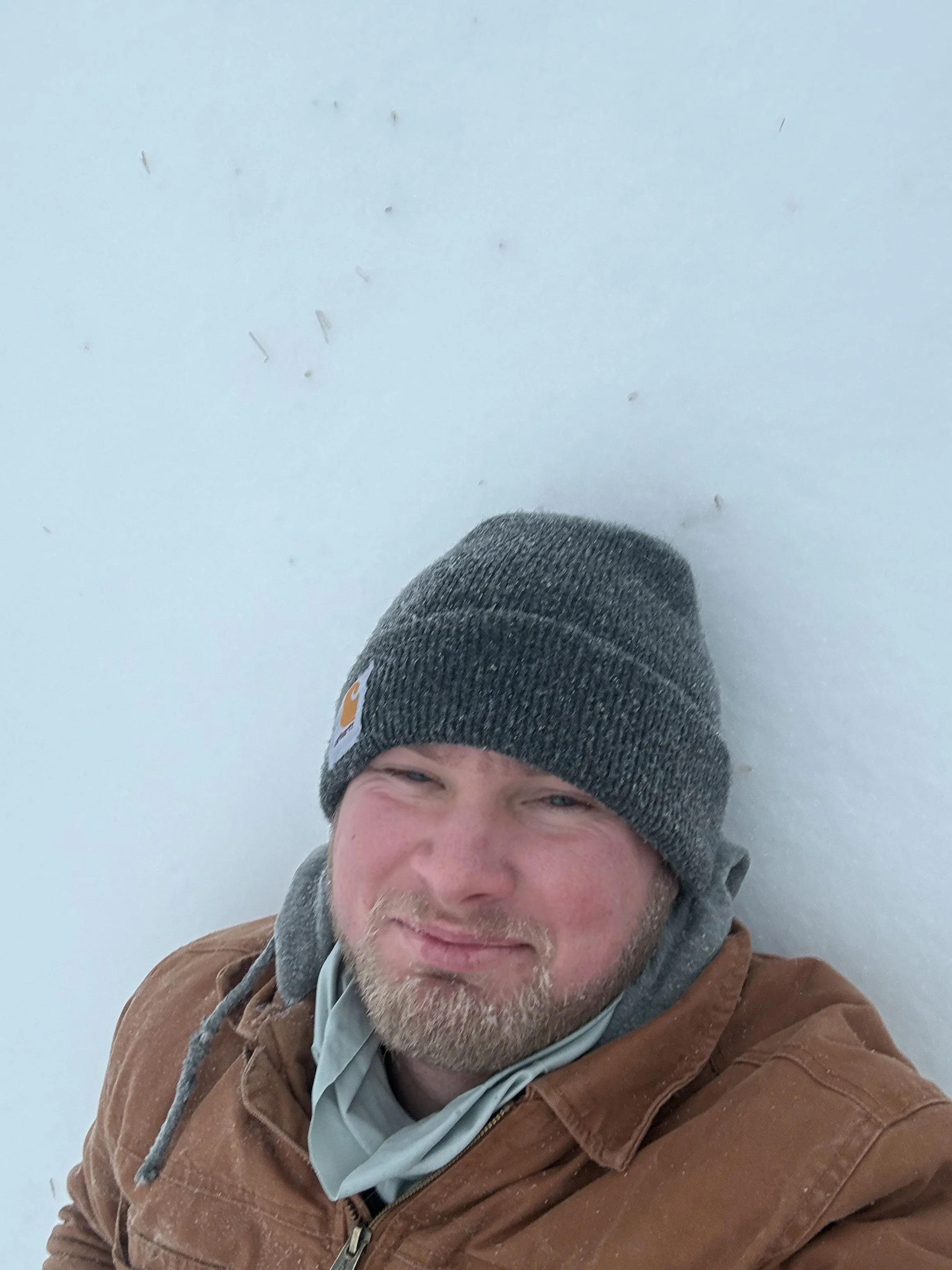 A man with a beard and mustache wearing a gray beanie, a light gray shirt, and a brown jacket, standing in the snow and smiling at the camera.