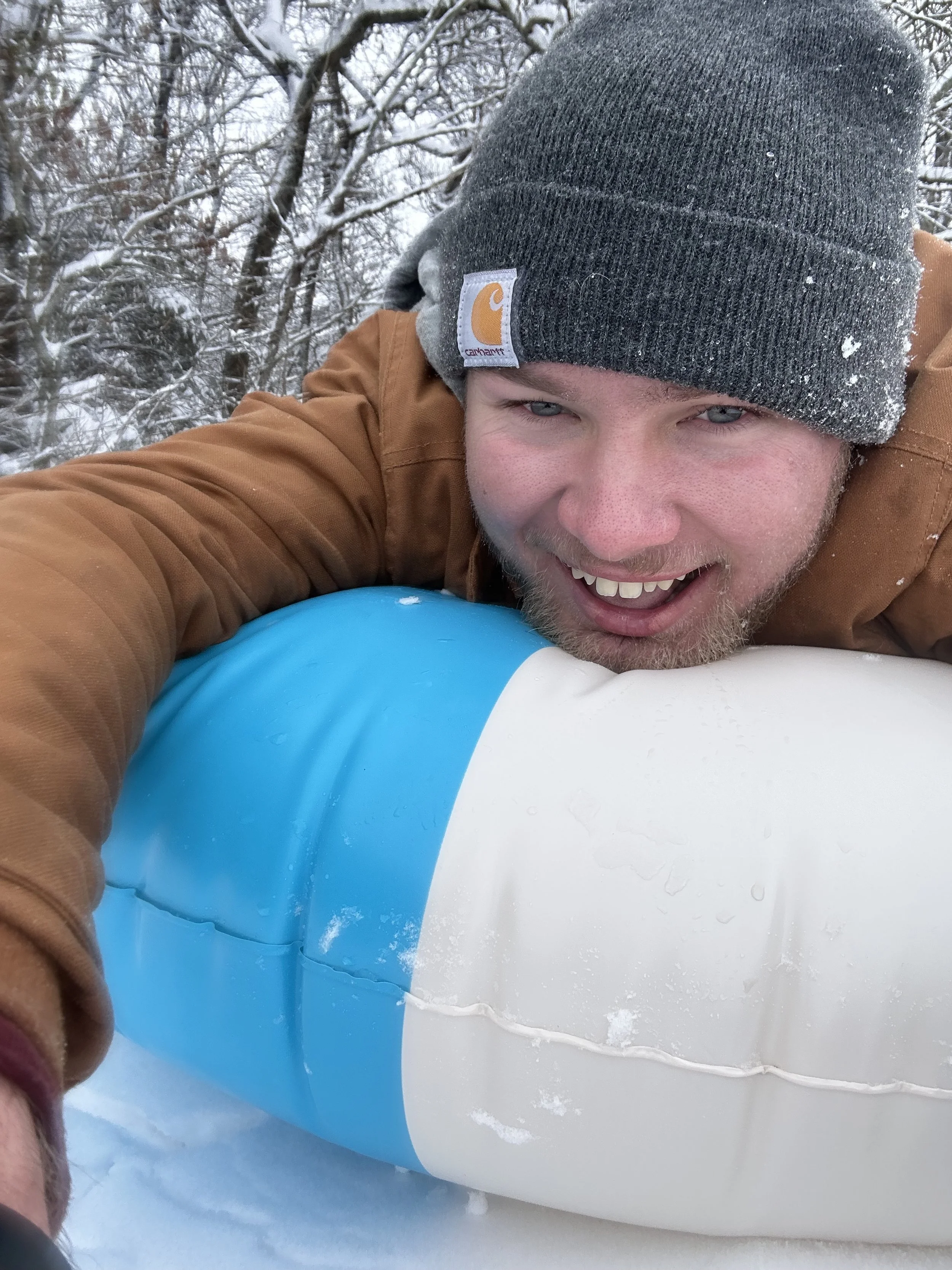 A man smiling, wearing a gray Carhartt beanie and a brown jacket, lying on a snow-covered surface with a blue and white object nearby, outside in a snowy wooded area.