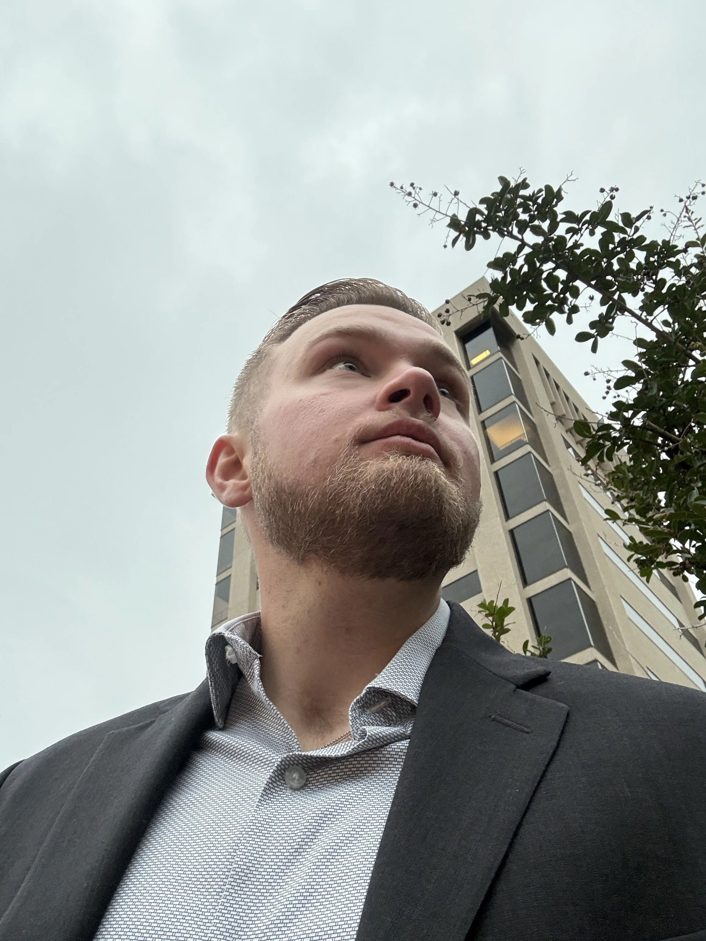 Portrait of a young man with short hair and a beard, wearing a dark suit and a light patterned shirt, taken outdoors with a building and trees in the background.