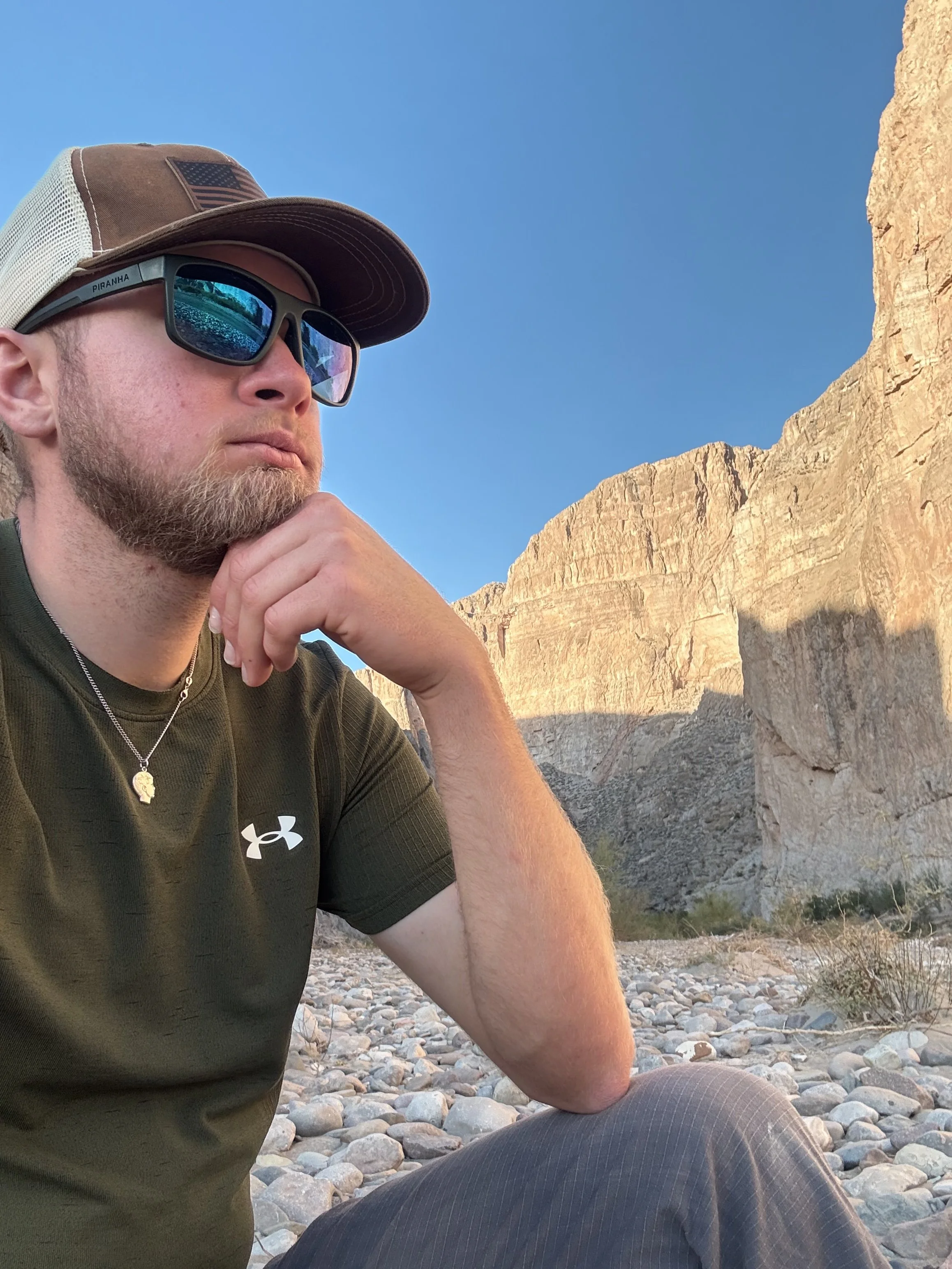 A man in sunglasses and a brown cap with an American flag patch, sitting outdoors with rocky terrain and large cliffs in the background.