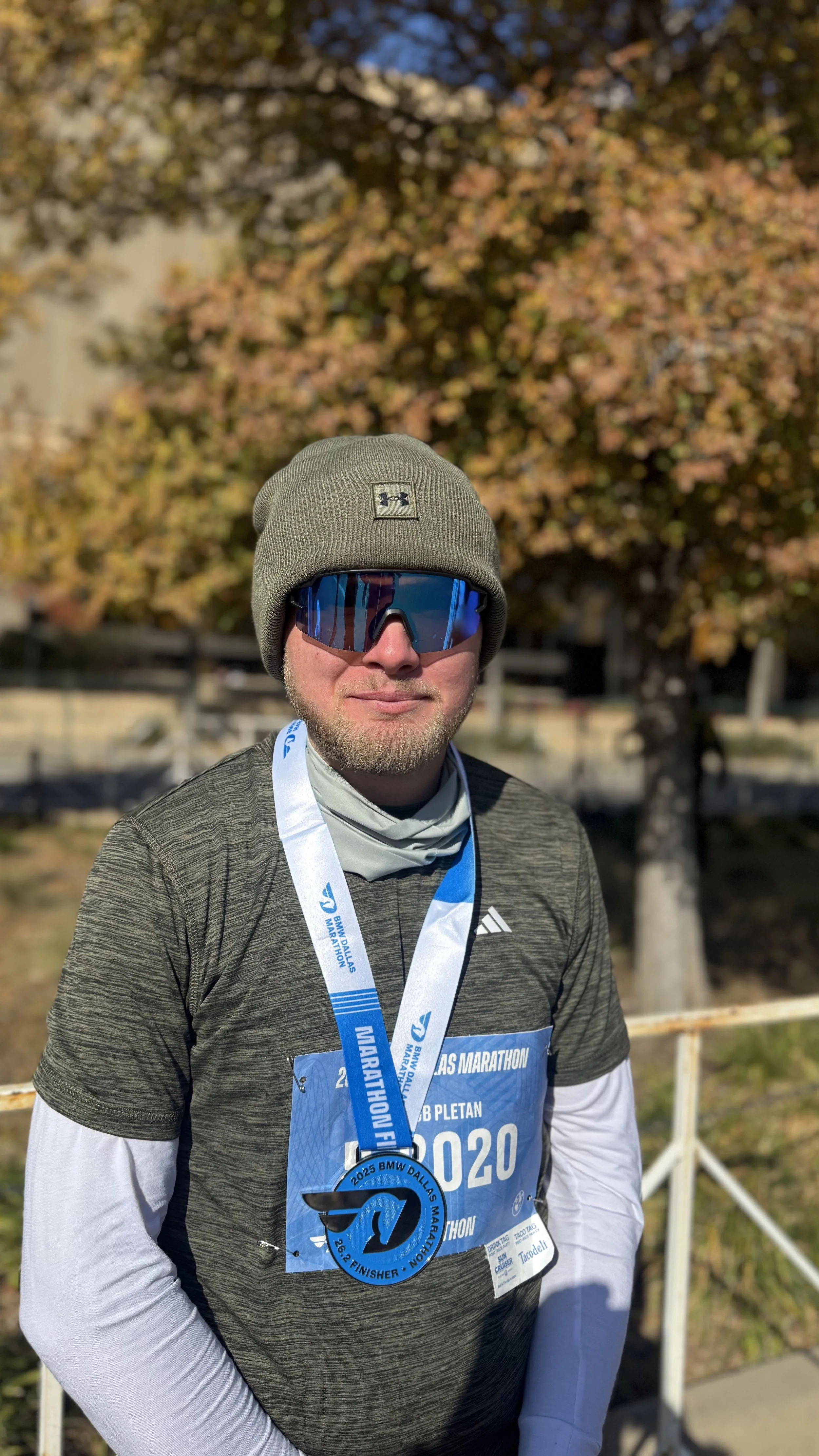 A man wearing sunglasses, a gray beanie, and a race bib with a finisher medal at a marathon event. Background features a tree with autumn-colored leaves.