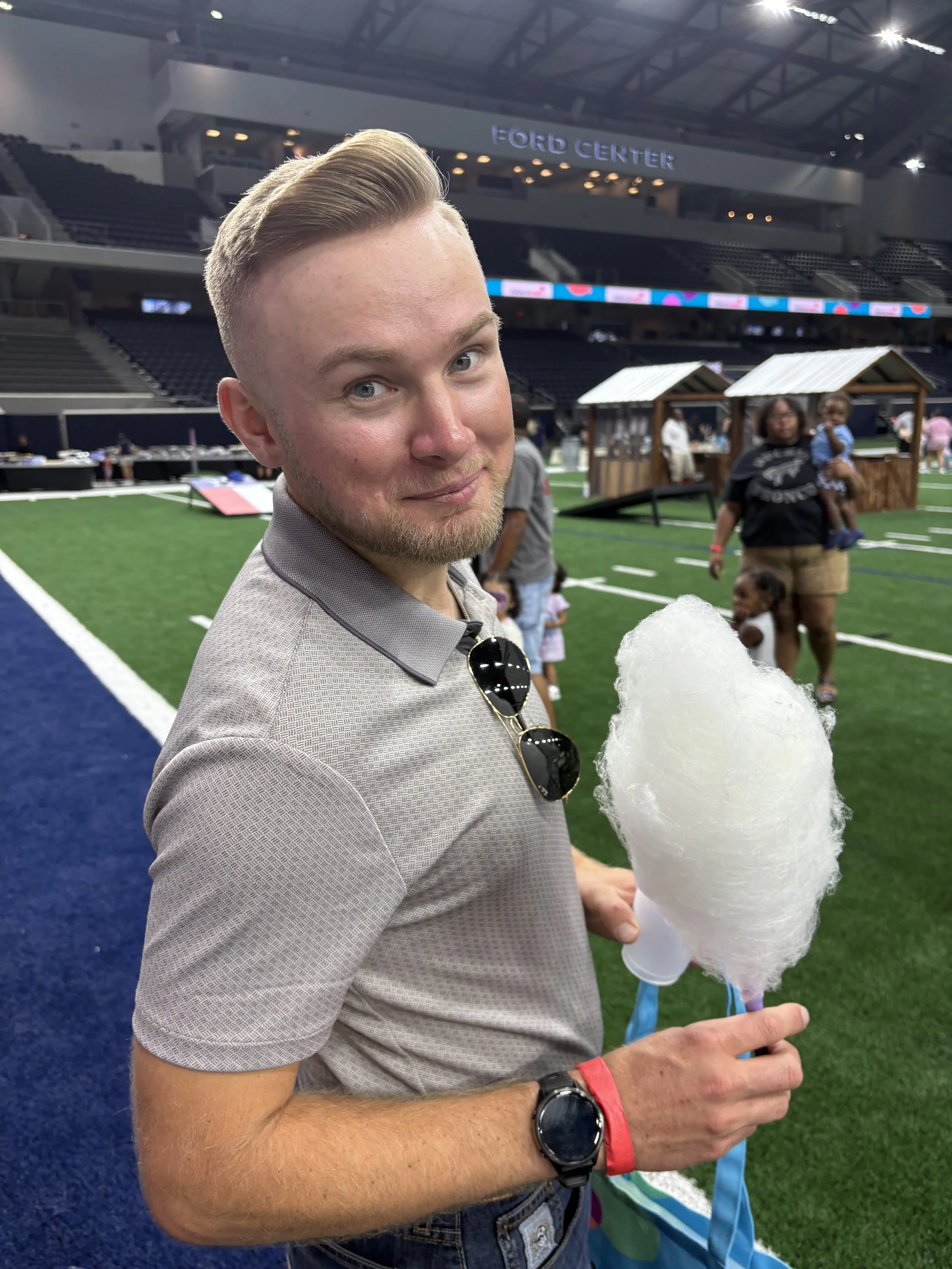 A man holding a cotton candy at an indoor sports arena with a crowd and a woman with a child in the background.