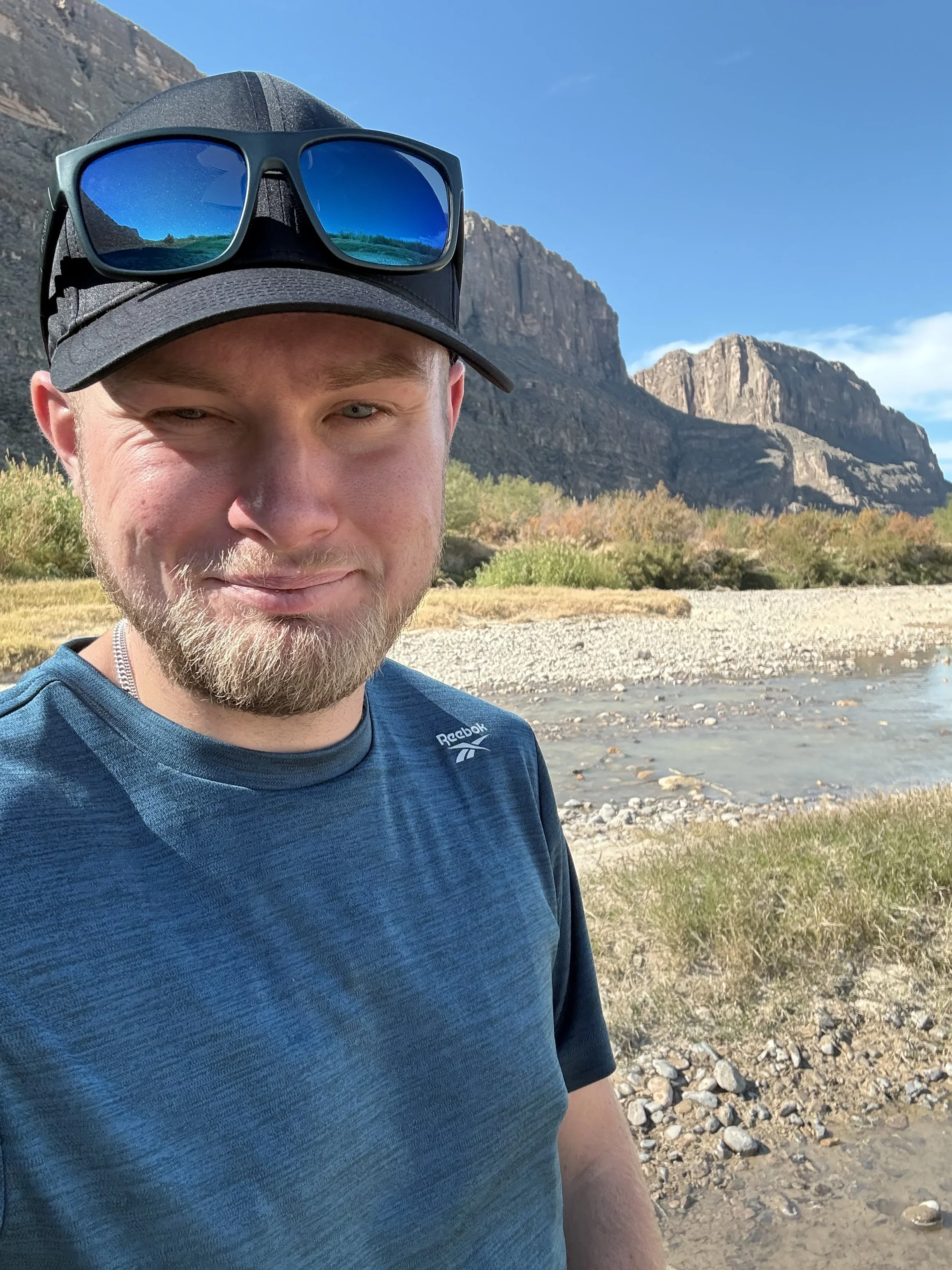A man in a blue Reebok shirt and gray cap with blue sunglasses on top, standing outdoors near a rocky riverbank under a blue sky with mountains in the background.