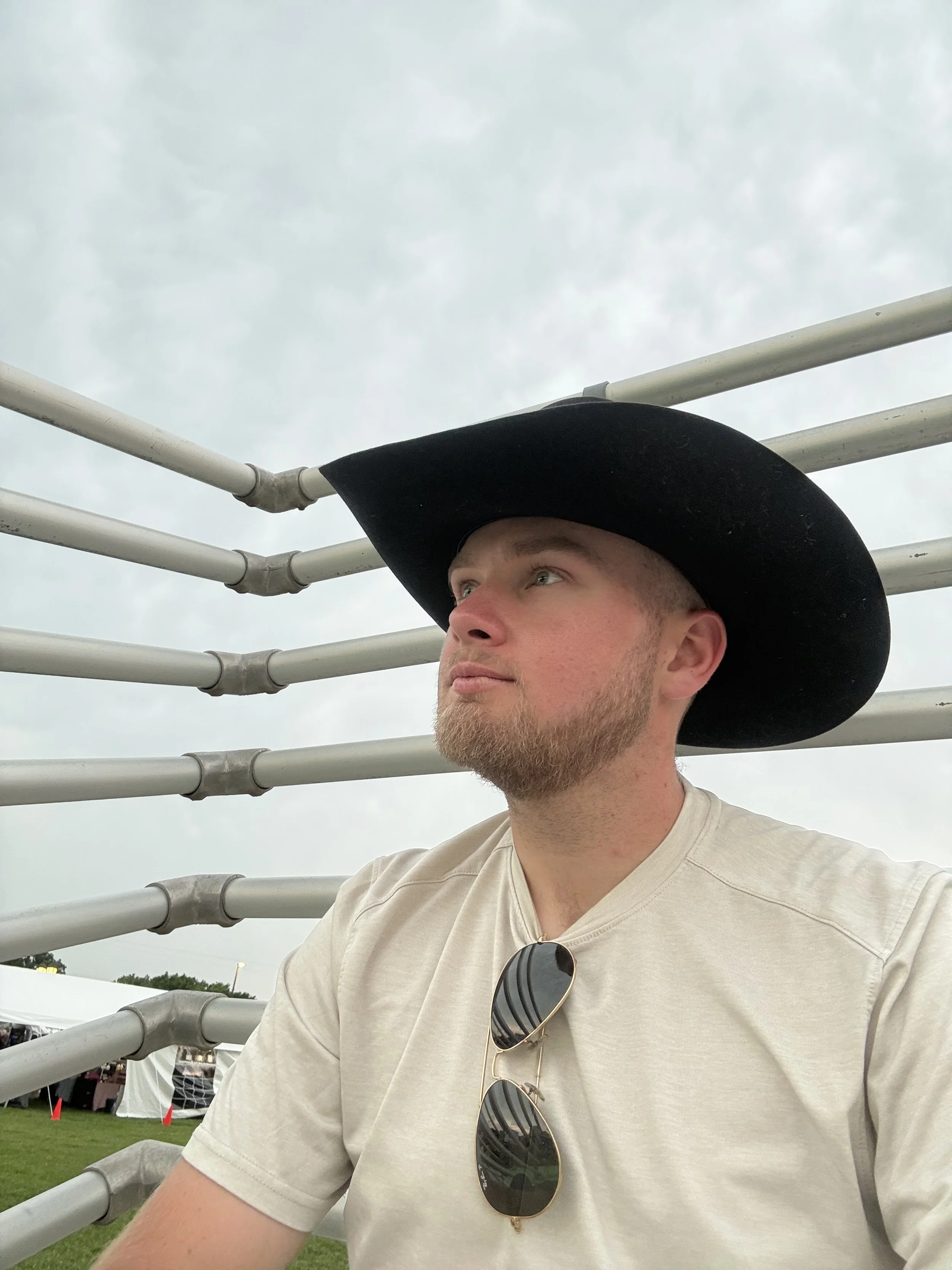A young man with a beard wearing a black wide-brimmed hat and sunglasses hanging from his beige T-shirt, sitting near metal bleachers outdoors on a cloudy day.