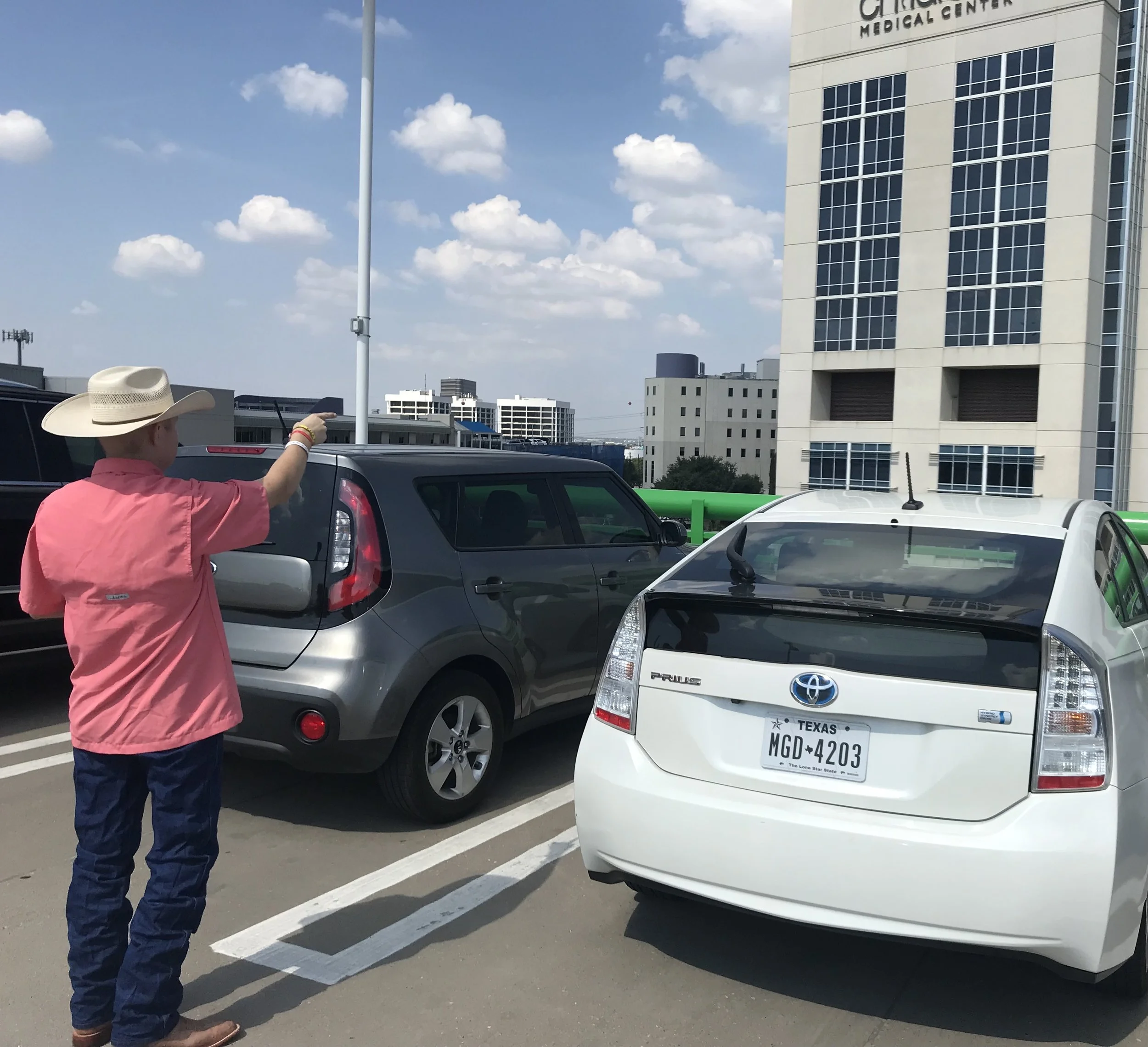 A woman with a cowboy hat and pink shirt appears to be pointing at a vehicle in a parking lot. The parking lot is partially filled with cars, and there is a large building with signs that read 'Children's Medical Center' in the background. The sky is