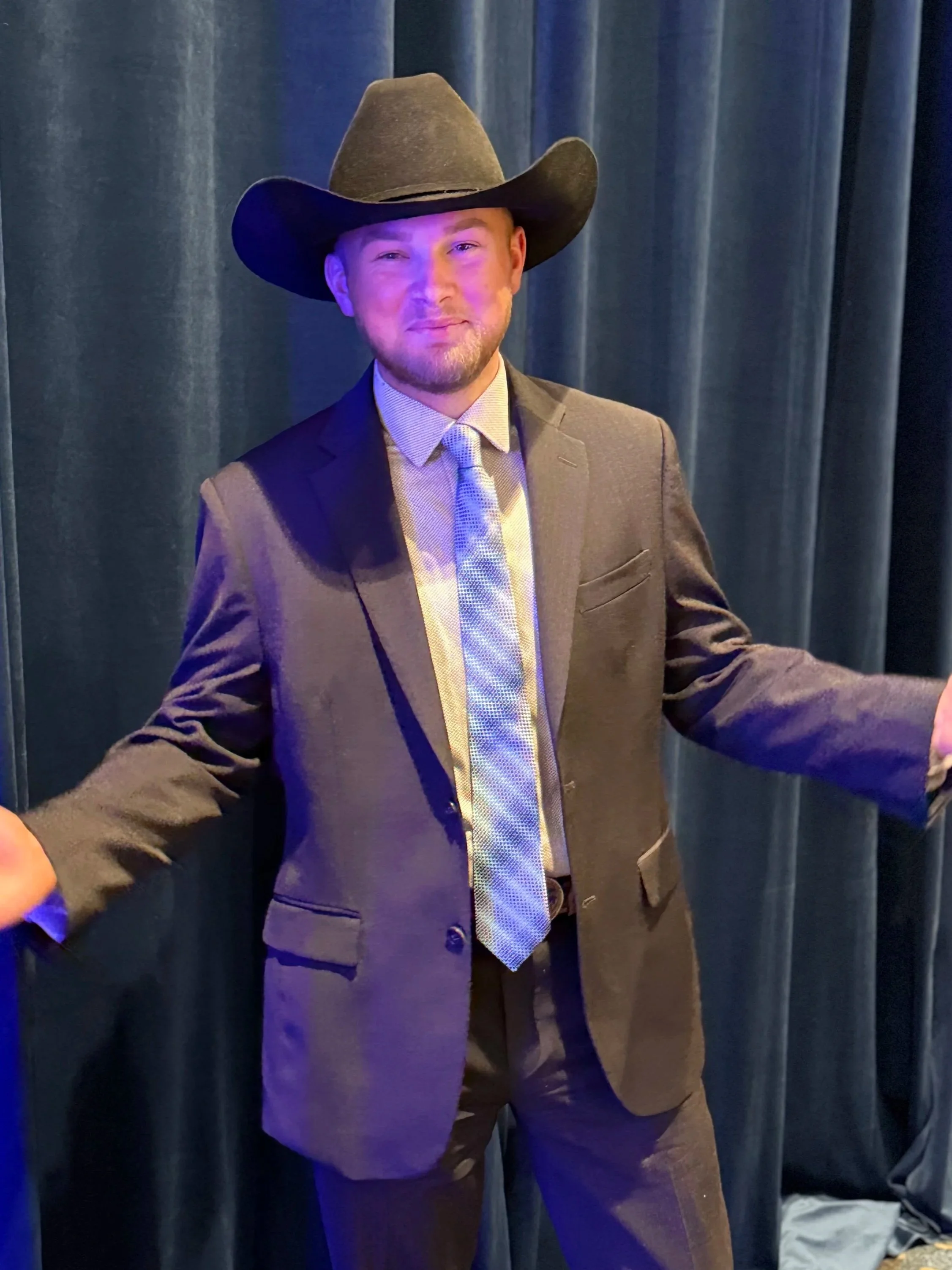 A man in a brown suit, patterned tie, and cowboy hat standing in front of dark curtains.