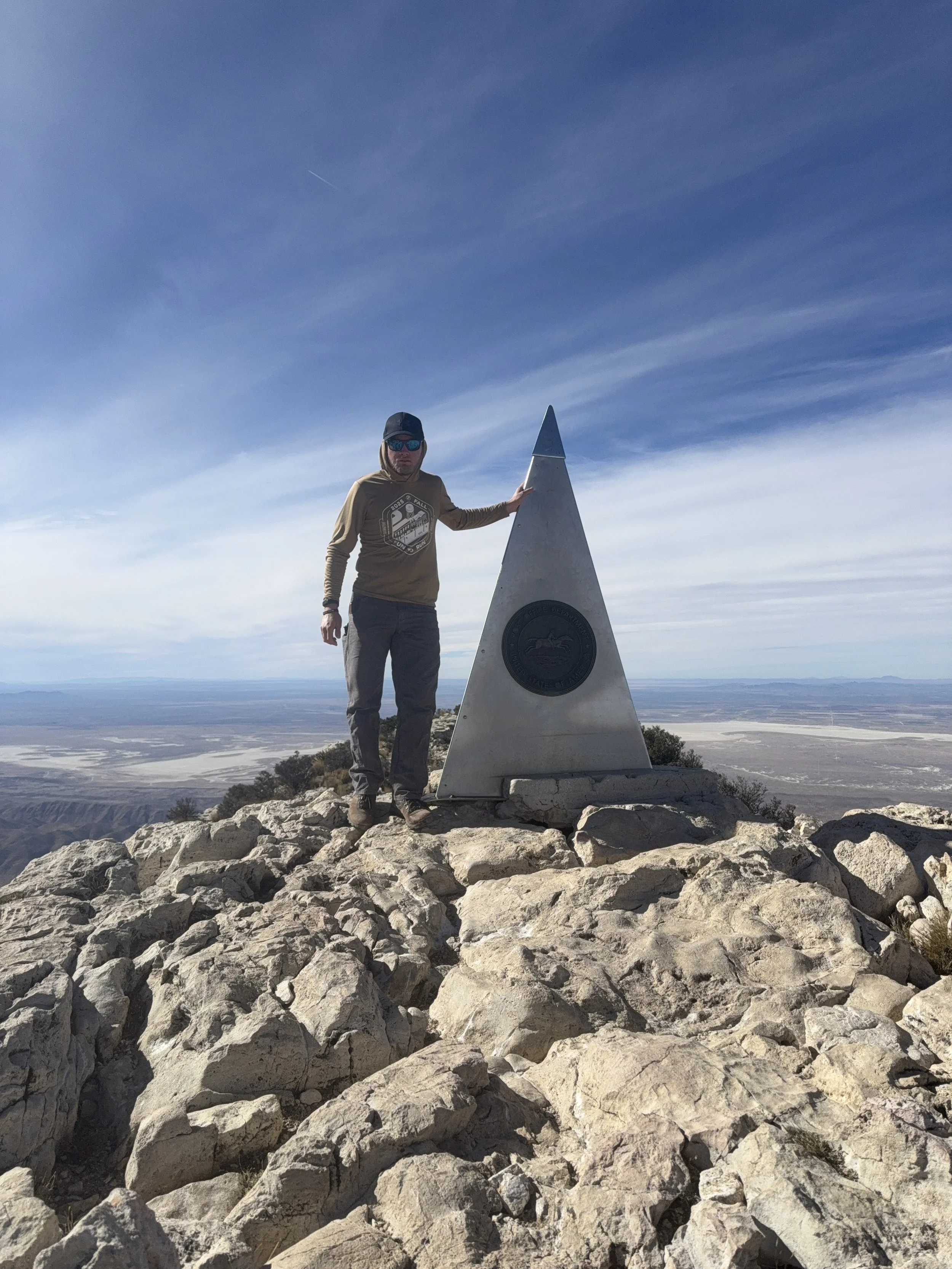 A man wearing sunglasses and a hat standing on rocky terrain next to a triangular summit marker with a seal, in a mountainous landscape with a flat valley and distant mountains under a blue sky.