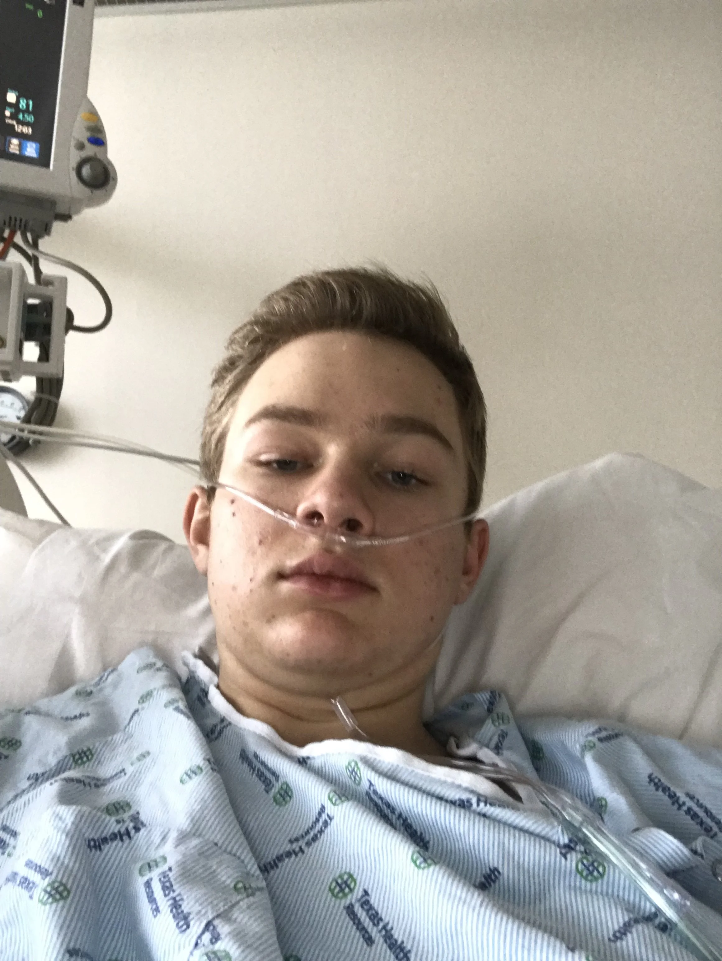 A young person with short hair and glasses lying in a hospital bed, connected to medical equipment and with an oxygen tube in their nose.