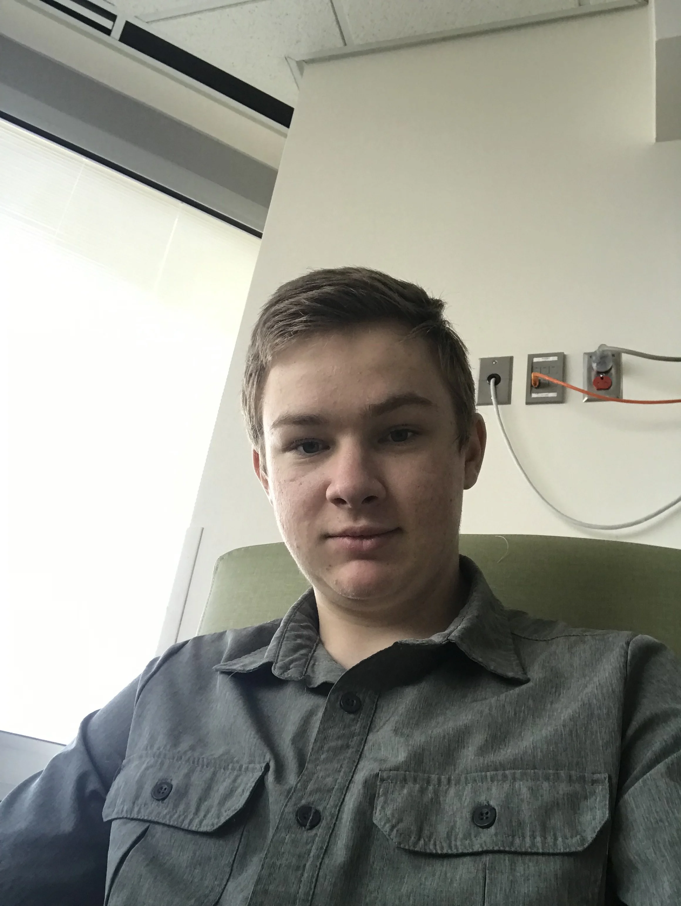 A young man with short brown hair and fair complexion taking a selfie in a room, sitting on a green chair, wearing a gray button-up shirt, with a white wall and electrical outlets in the background.