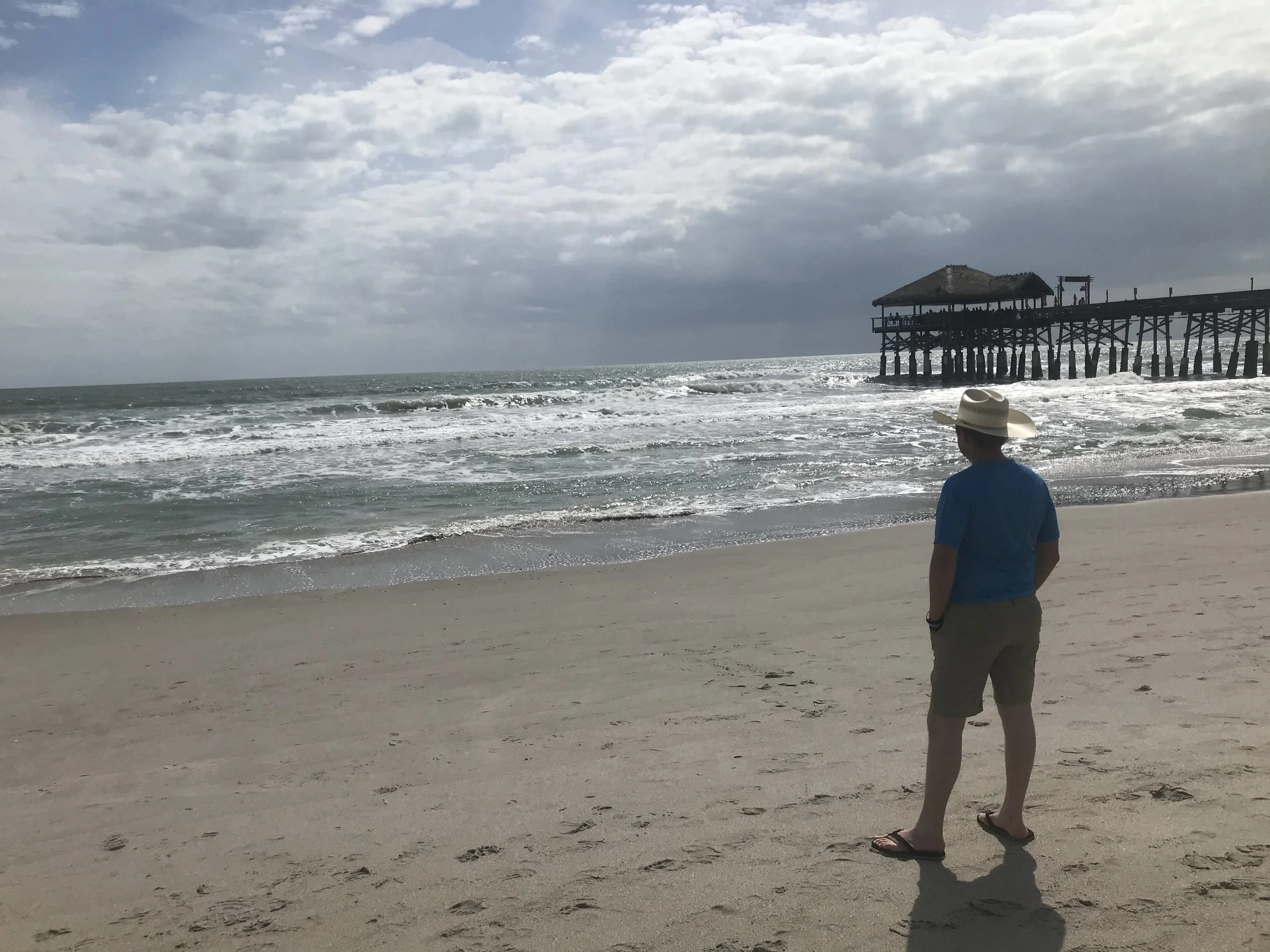 A person standing on a sandy beach, facing the ocean with small waves, under a cloudy sky. The person is wearing a straw hat, a blue shirt, and shorts, with their hands in their pockets. In the background, there is a wooden pier with a thatched-roof 