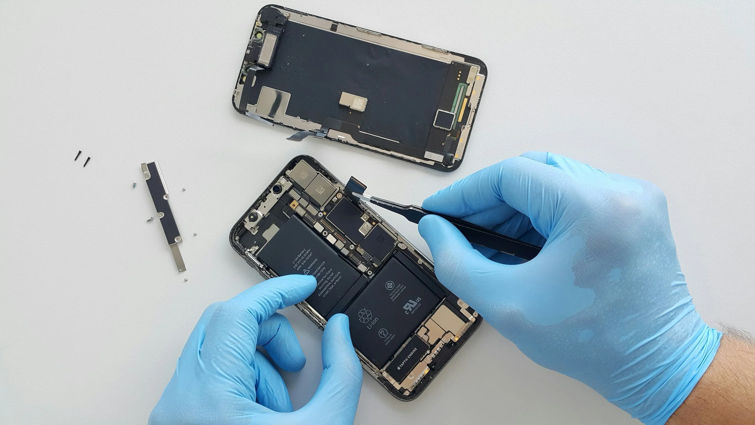 A person wearing blue gloves is repairing a disassembled smartphone, holding the phone with one hand and using a tool with the other, with the phone's internal components exposed on a white surface.