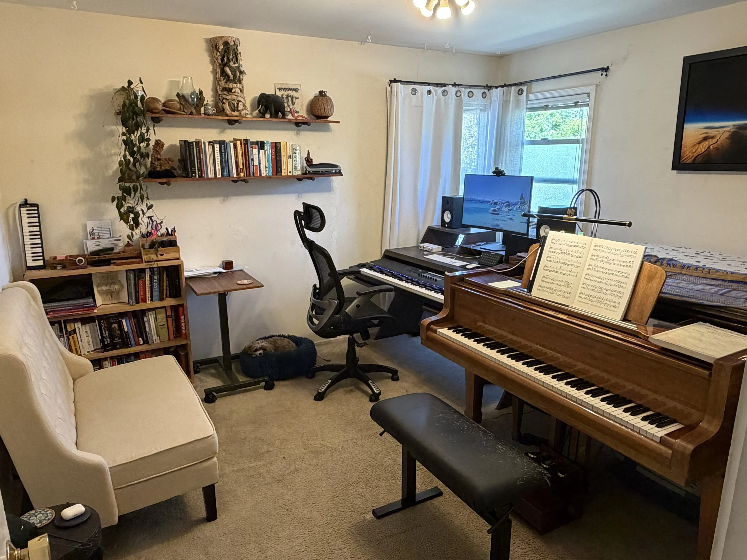 John Metz's piano studio, with a wood grand piano, a digital keyboard workstation, shelves of music books, and a small dog napping in its bed.