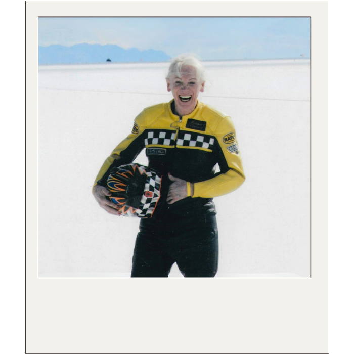 Polaroid of Connie Beavers, smiling woman in yellow and black car racing outfit, holding helmet.