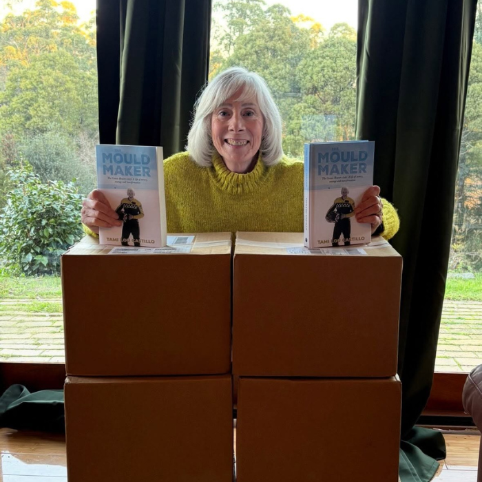Tami Lou Castillo holding two copies of her book The Mould Maker. She's behind two stacks of cardboard boxes and she's holding her books up in top of the boxes.