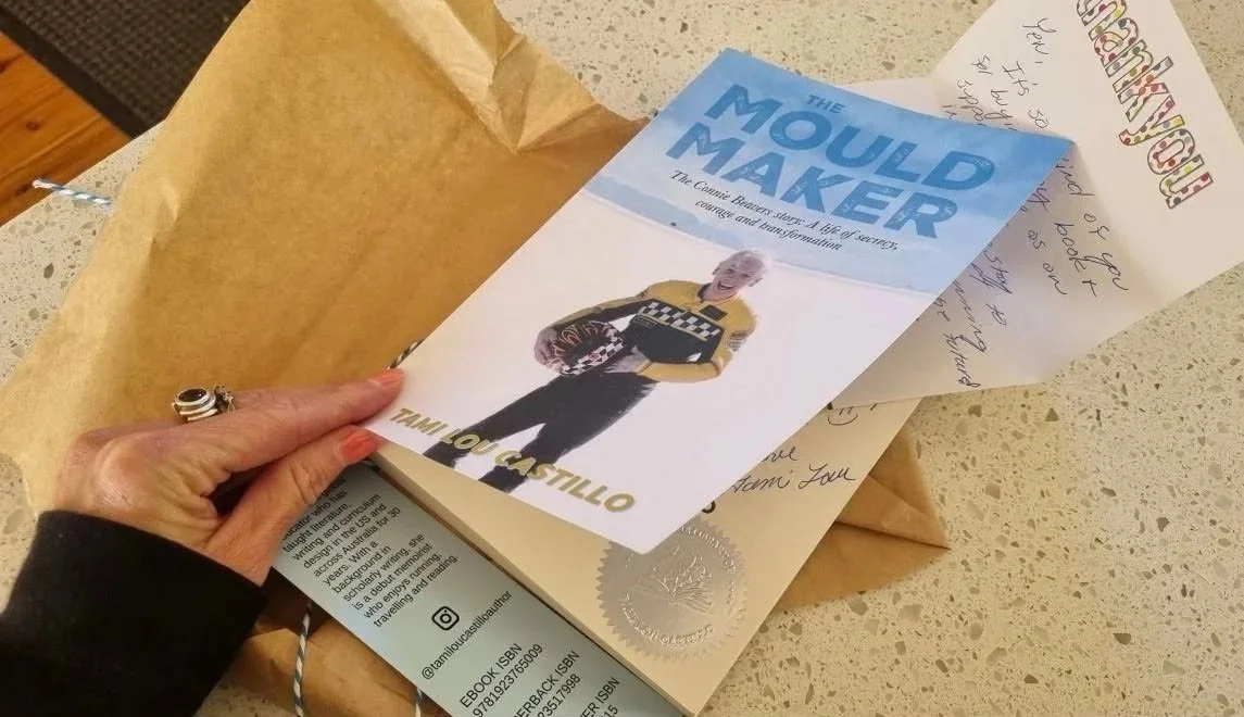 Hand opening their unwrapped, signed copy of The Mould Maker by Tami Lou Castillo, with a thank you card peeking out