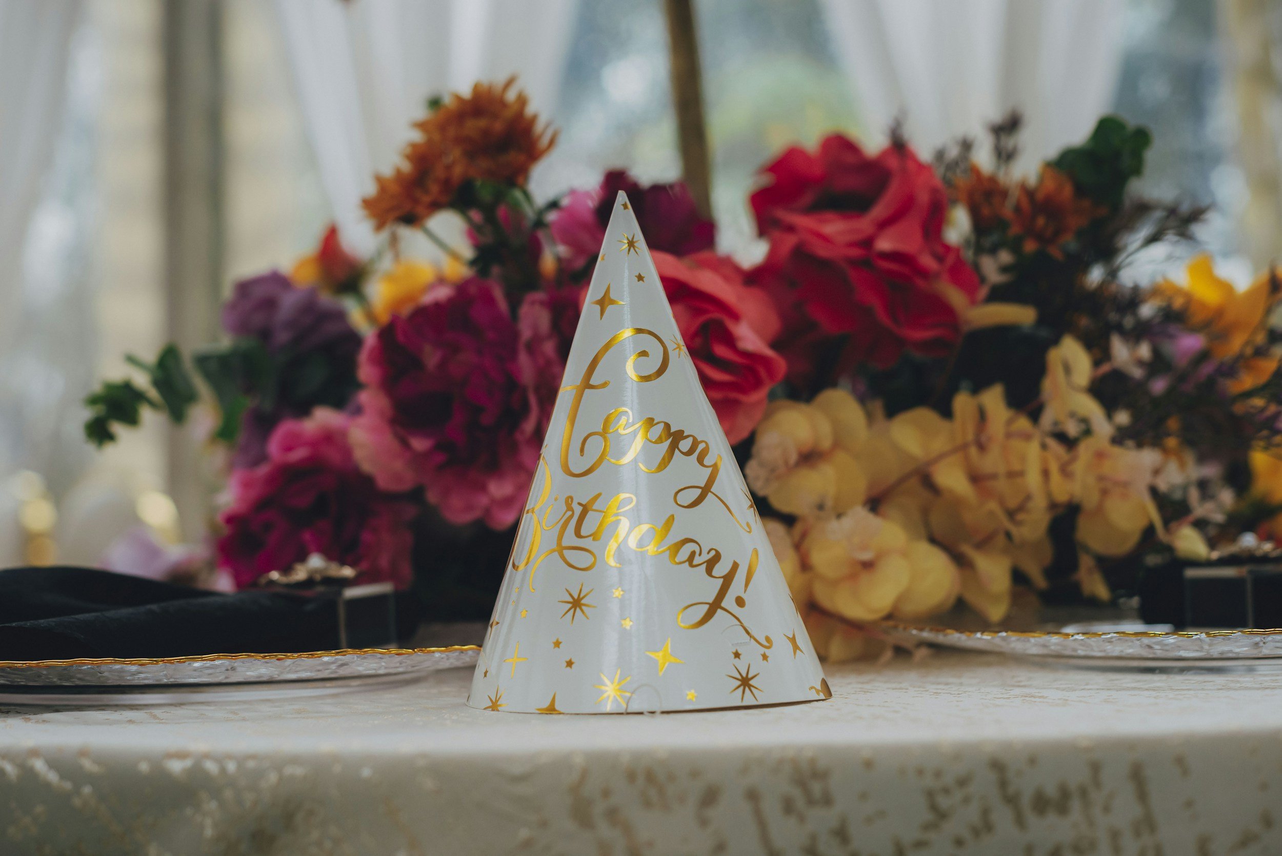 Celebration hat with 'Happy Birthday!' written on it, placed on a table in front of a colorful flower arrangement.