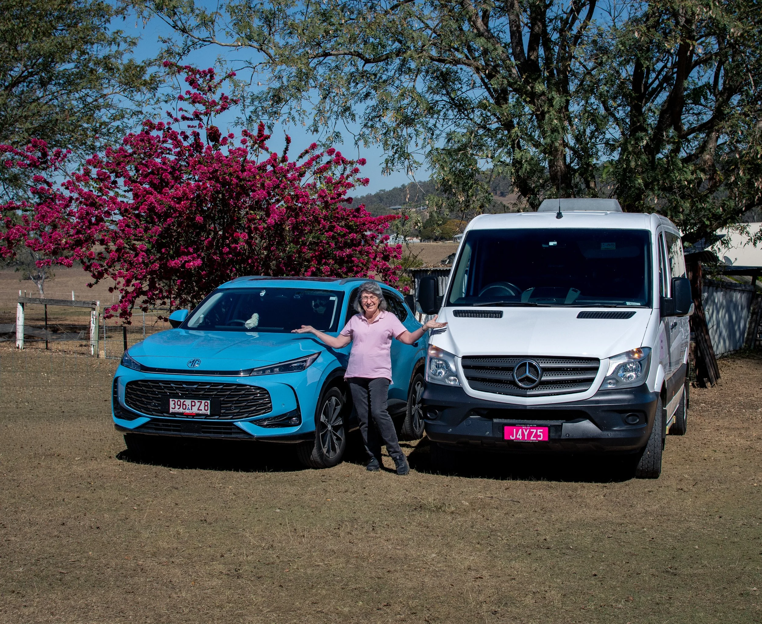 A woman in pink standing between a blue car and a white van in an outdoor rural setting with trees, pink flowering bushes, and a fenced area behind her.
