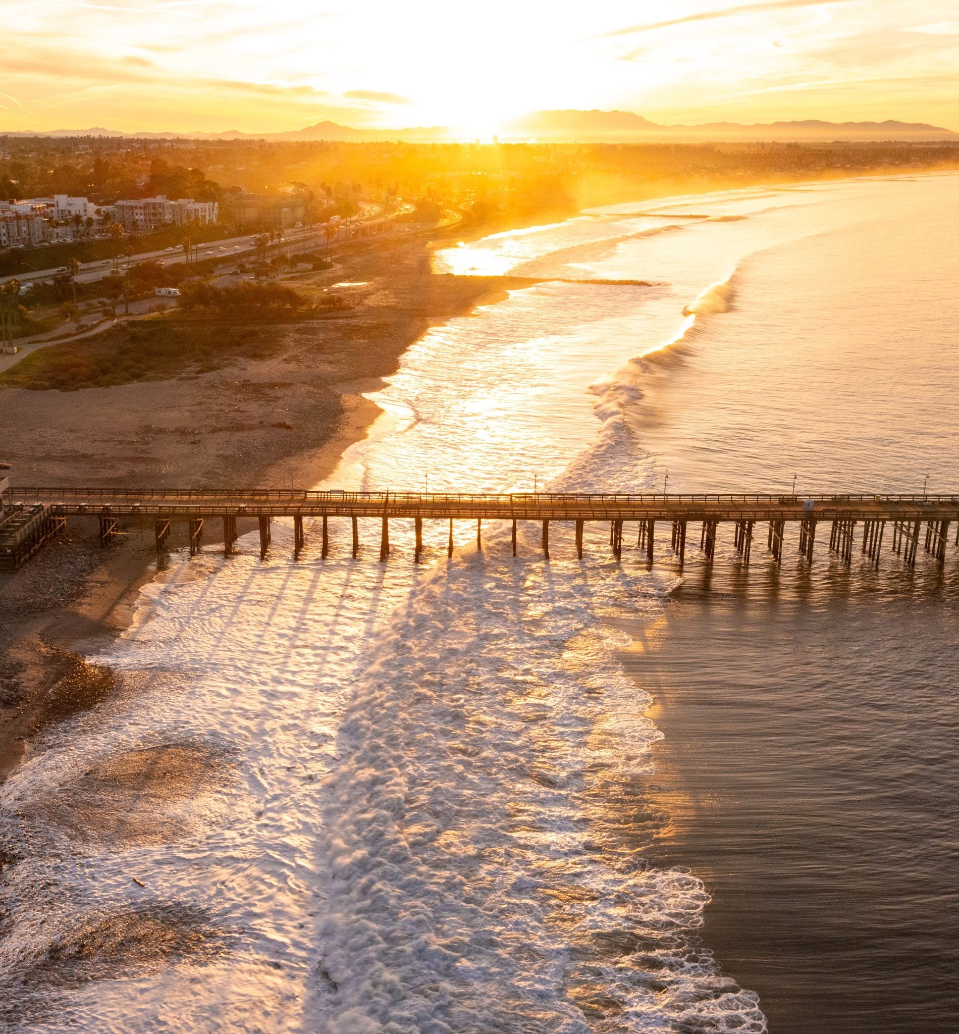 Sunset over the ocean with waves crashing on the shore and a coastal city in the distance.