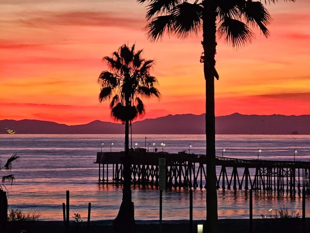 Sunset over a calm ocean with a pier extending into the water, tall palm trees in the foreground, and distant mountain silhouettes on the horizon.