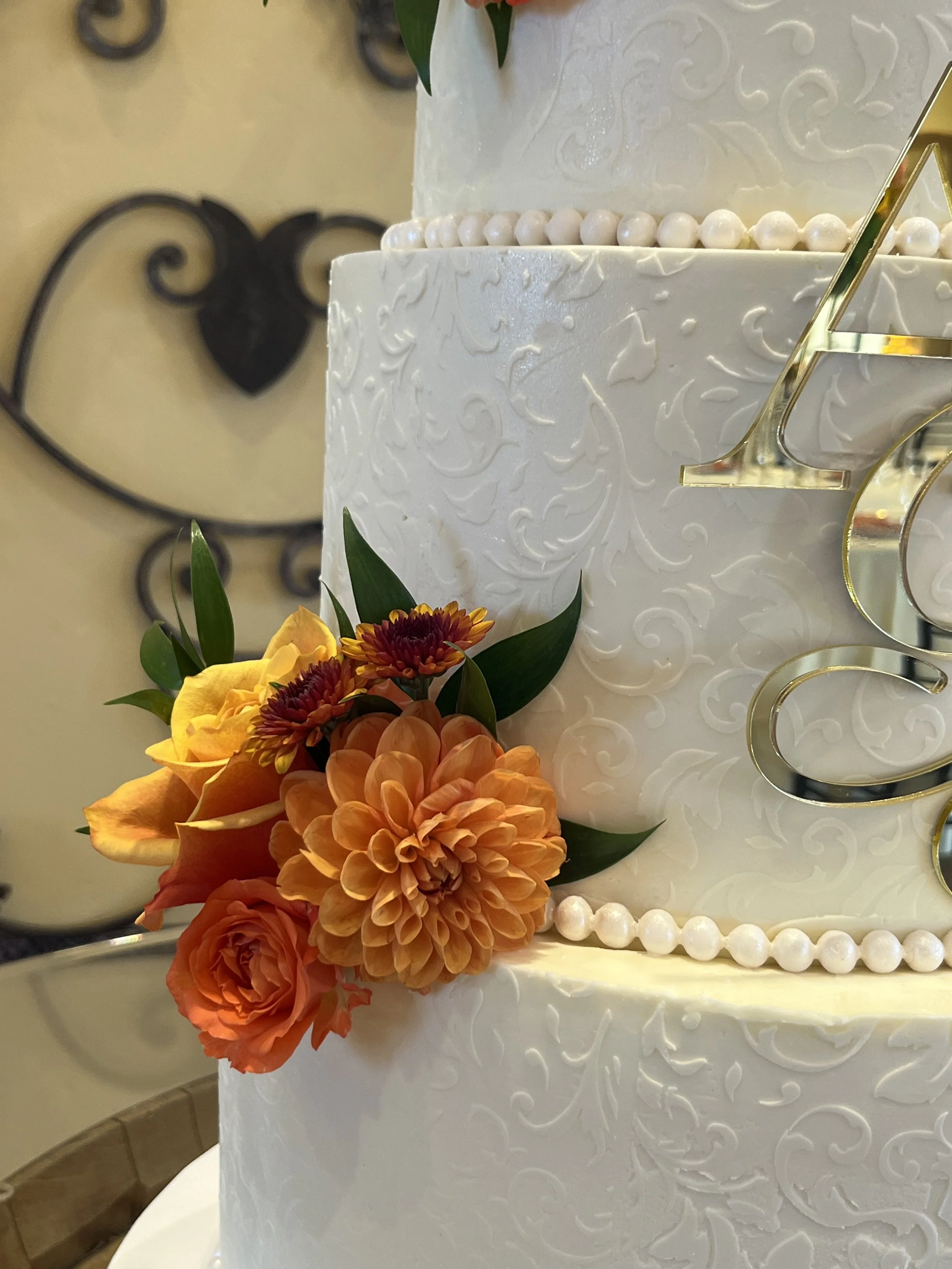 A white wedding cake decorated with embossed scroll and leaf designs, with a string of small white pearls around the top edge, topped with a gold 'D' letter. The cake is adorned with colorful fresh flowers including orange and yellow blossoms and gre