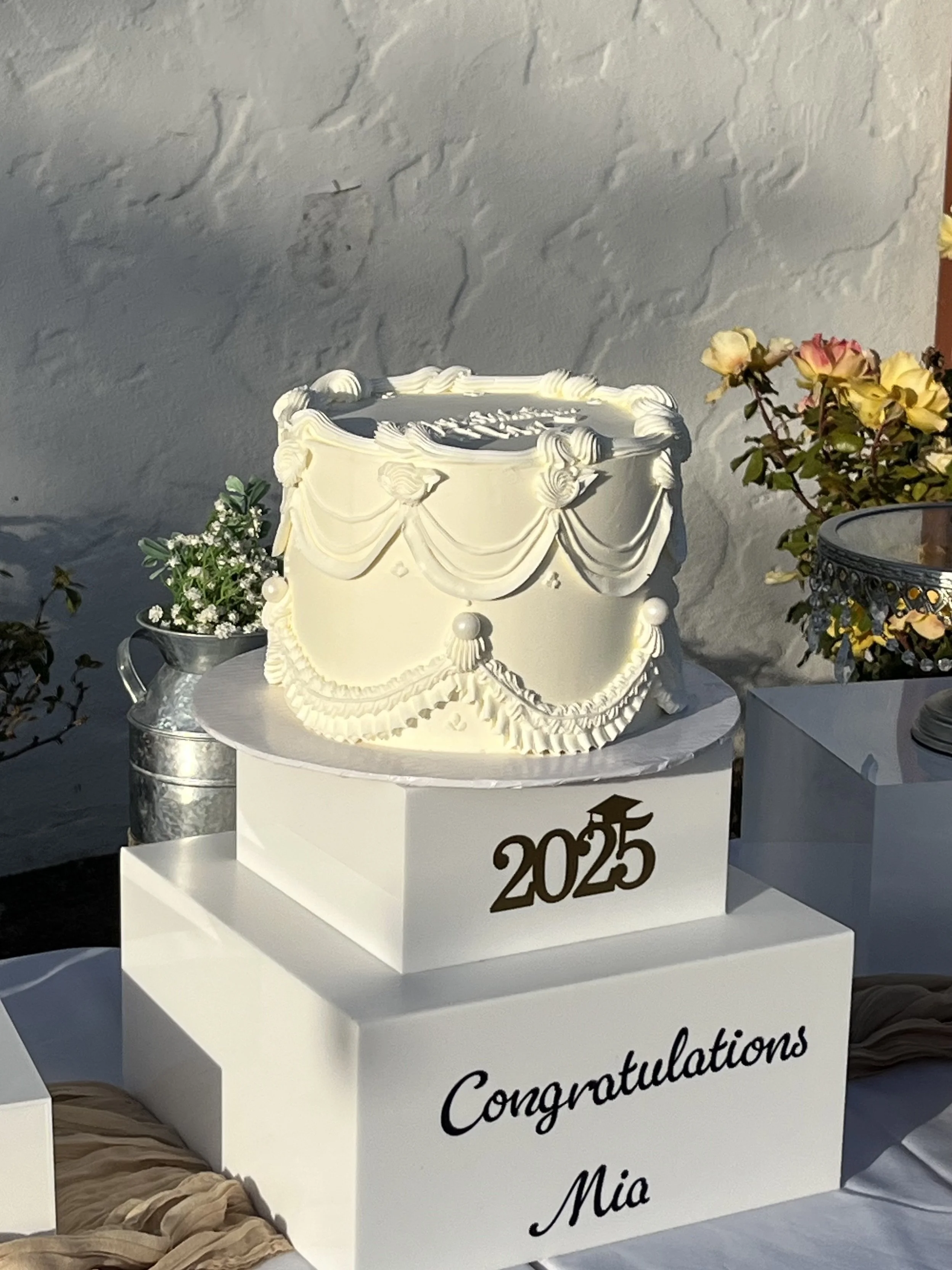 A white winner's cake with decorative piping and a small flower, placed on a white table, with a sign that says '2025 Congratulations'. There are flowers and a metal container nearby.