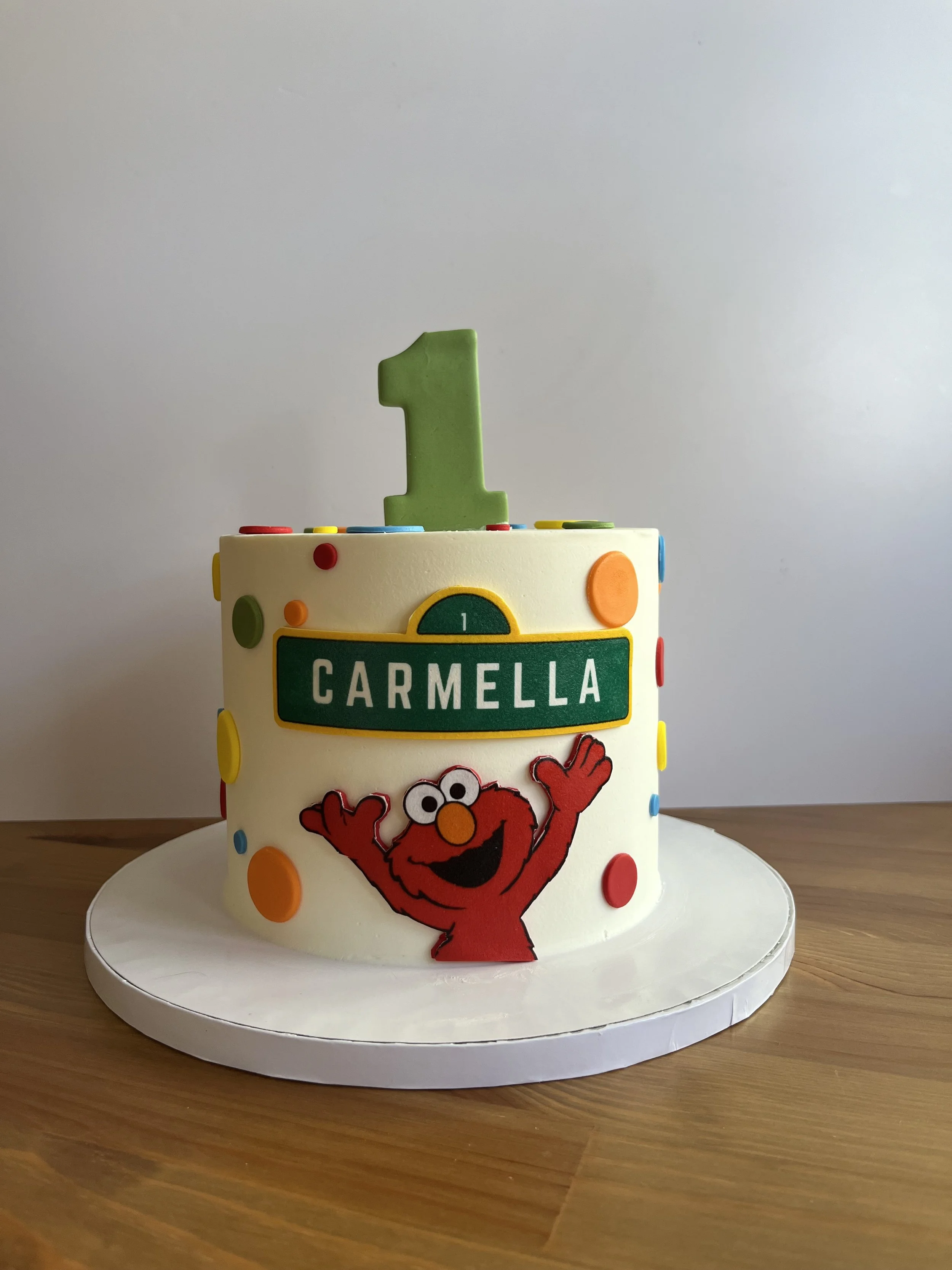 A birthday cake decorated with colorful dots, featuring a picture of Elmo from Sesame Street, and with a green number one candle on top.