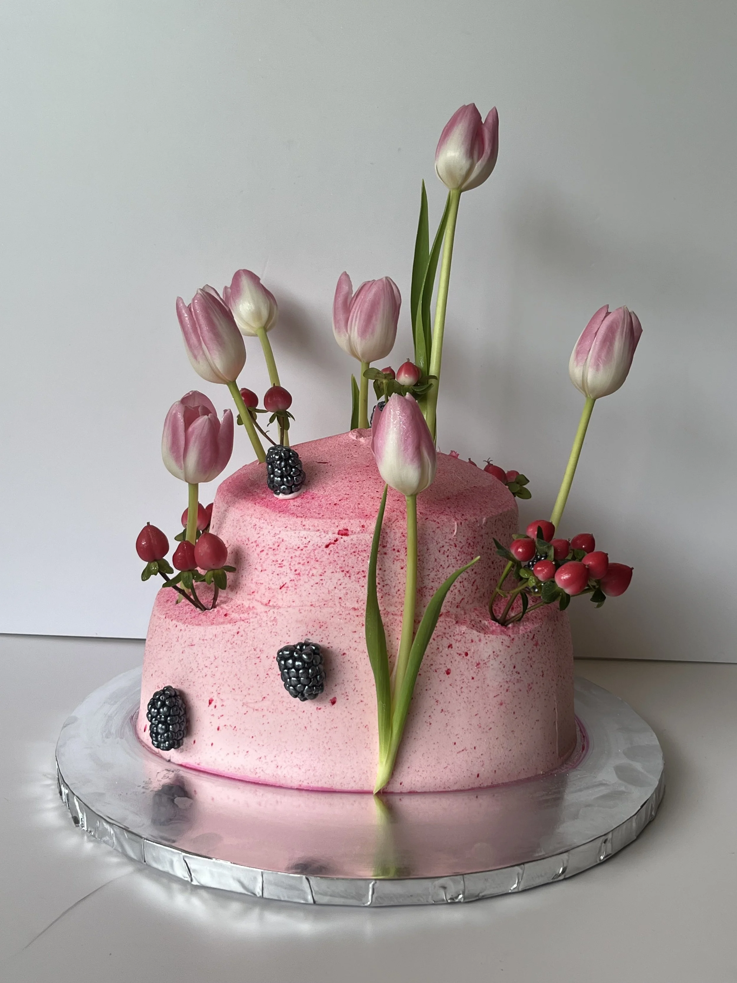 Pink two-tiered cake decorated with pink tulip flowers, blackberries, and red berries, sitting on a silver cake board.