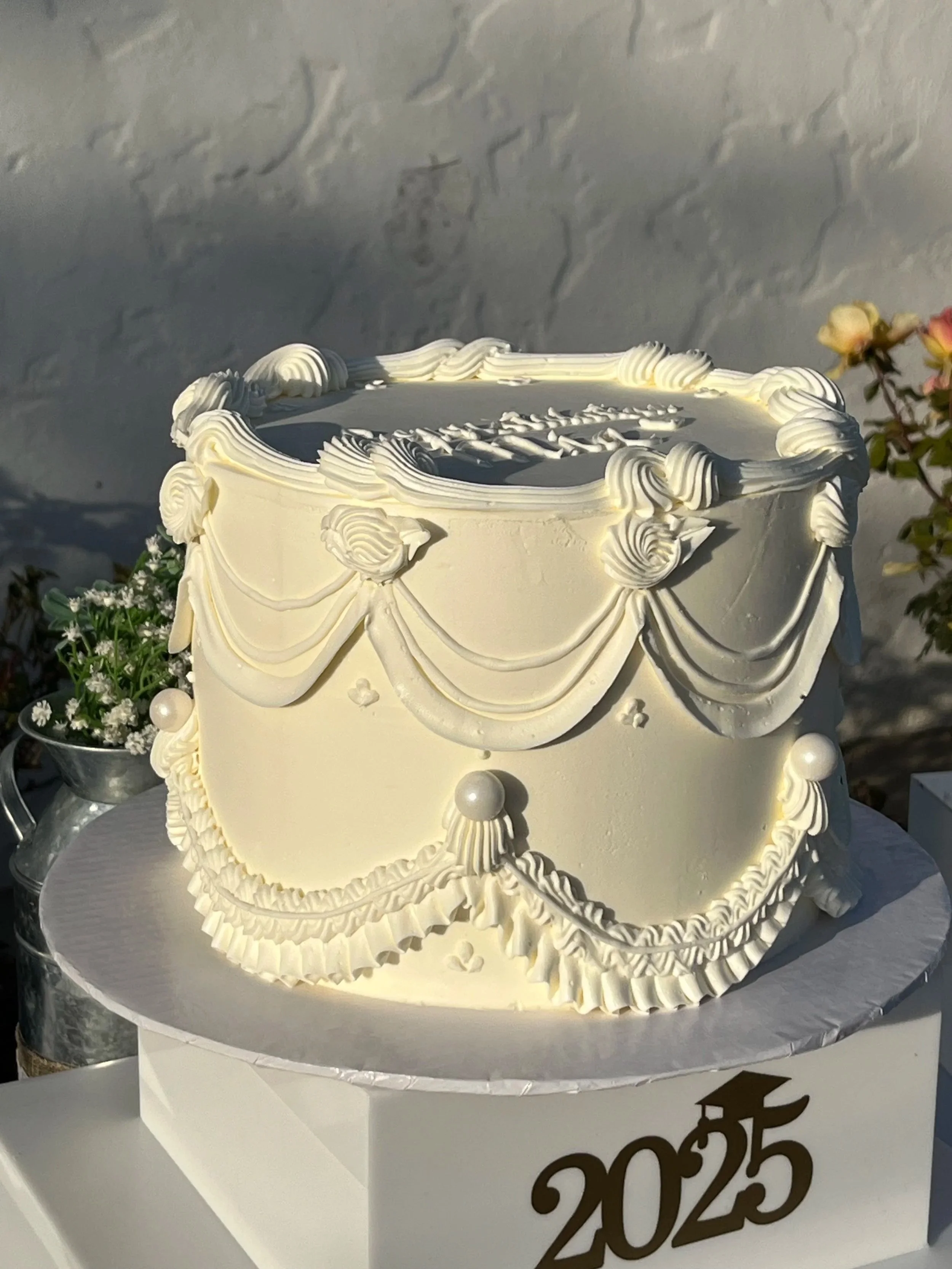A white cake decorated with piped icing and a '2023' cake topper, sitting on a white cake stand. Flowers are visible in the background.