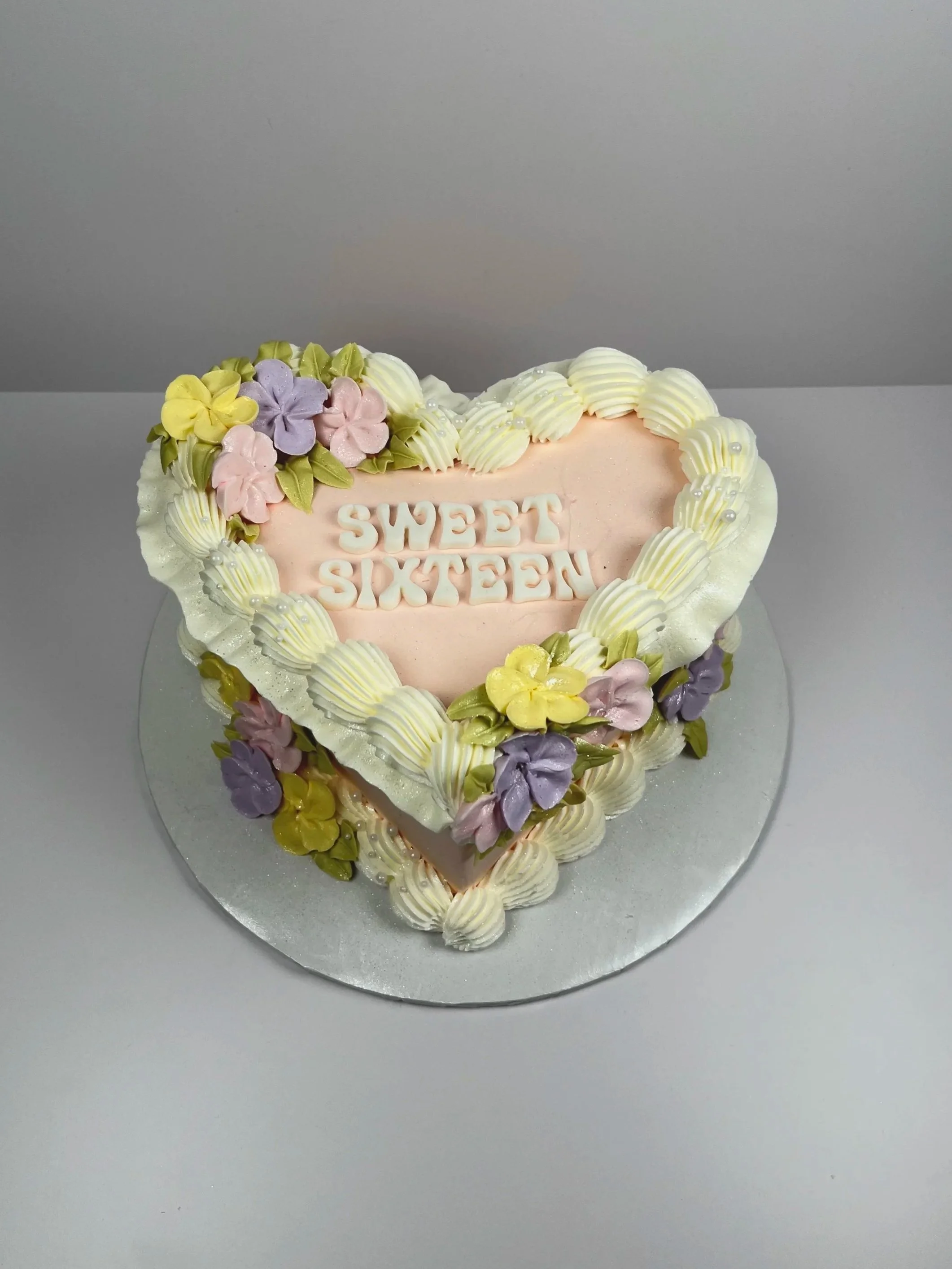 Heart-shaped cake with white and pastel pink, yellow, and purple flowers, and white piped decorations, with the words 'Sweet Sixteen' on top.