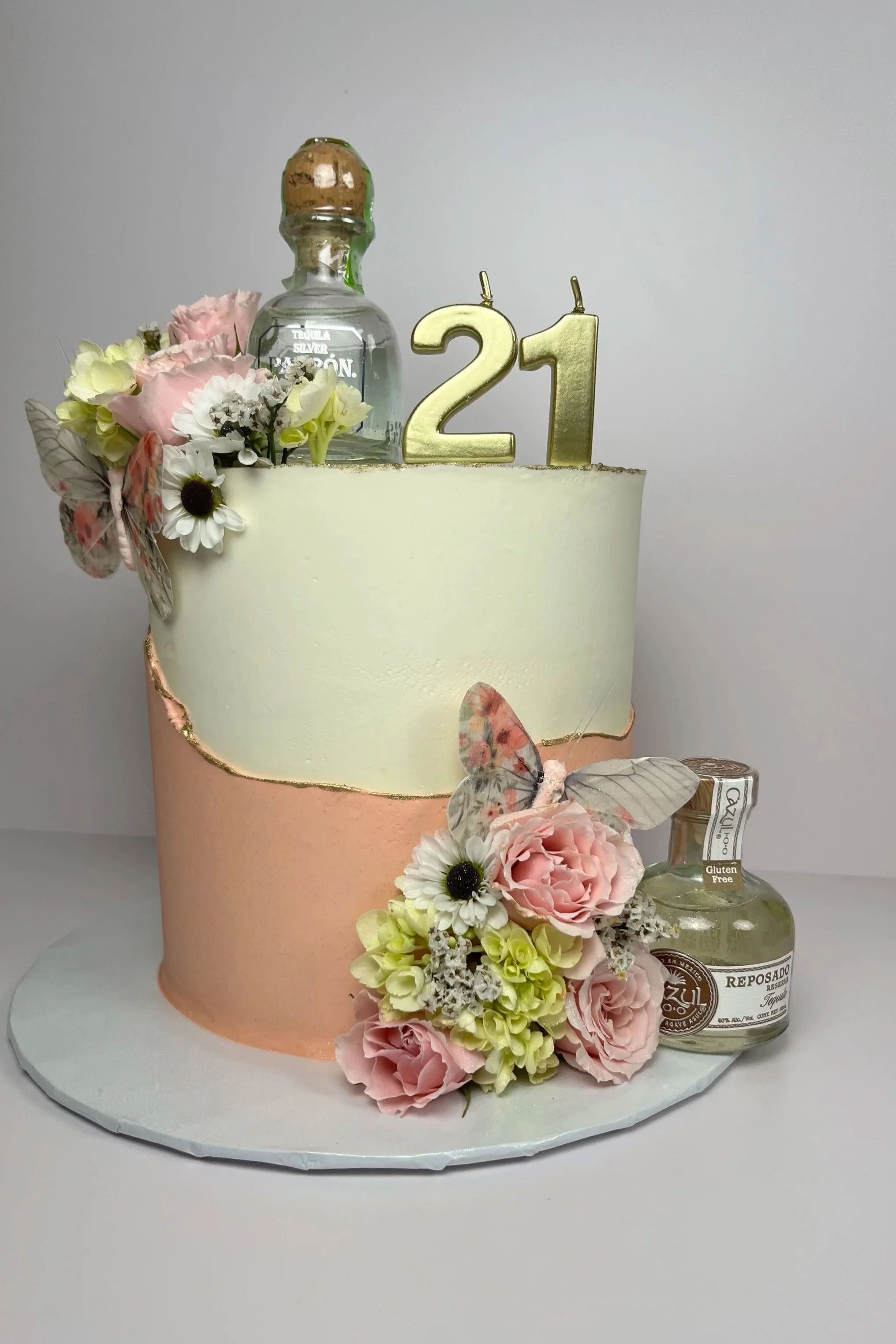A two-tone birthday cake decorated with pink and white flowers, a gold "21" candle, a clear liquor bottle, and a small bottle of tequila, all on a white cake board.