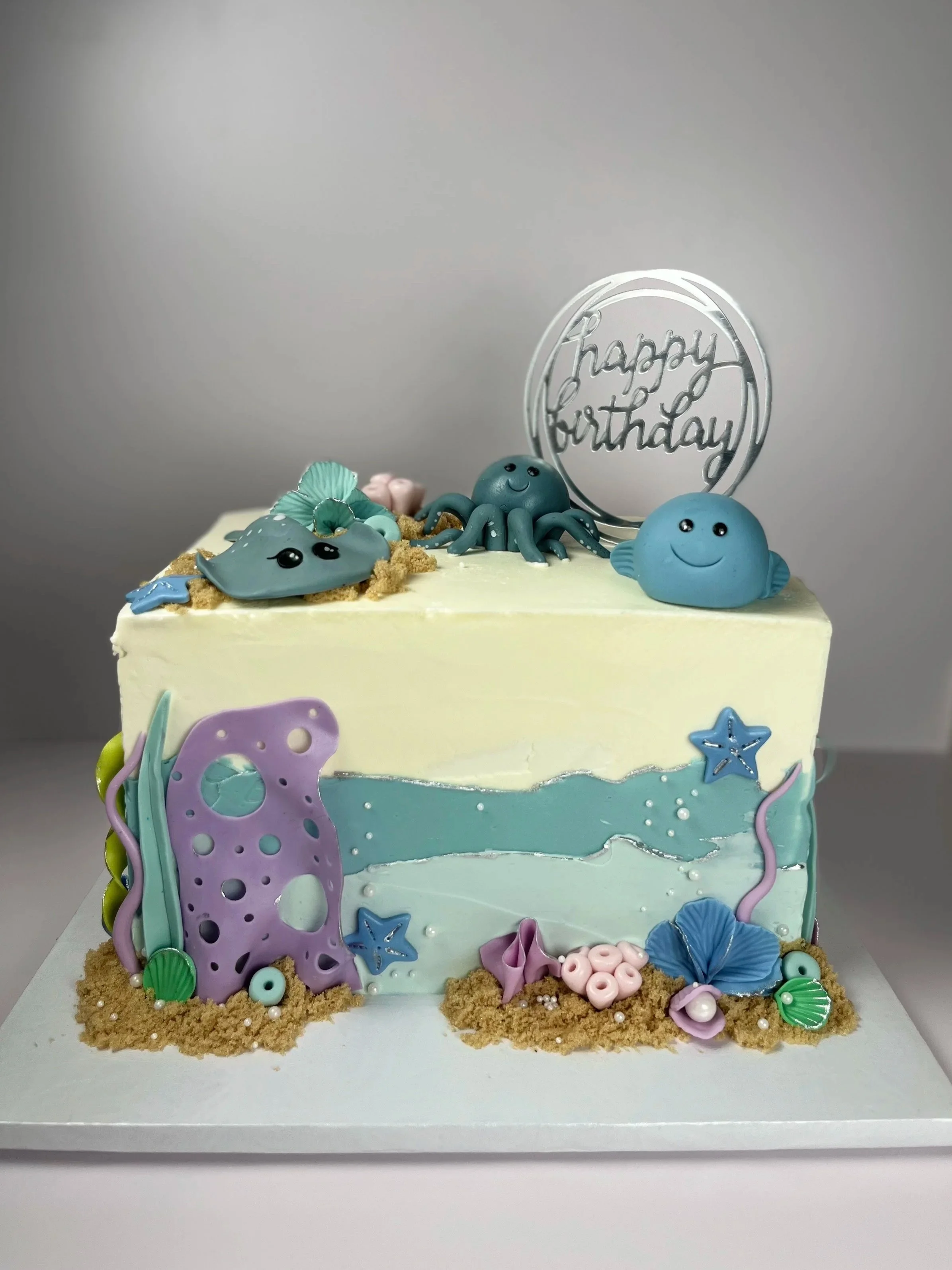 A colorful birthday cake decorated with ocean-themed fondant figures including a smiling octopus, fish, starfish, shells, and other sea creatures, with a silver "happy birthday" topper and sandy-colored decorations at the base.