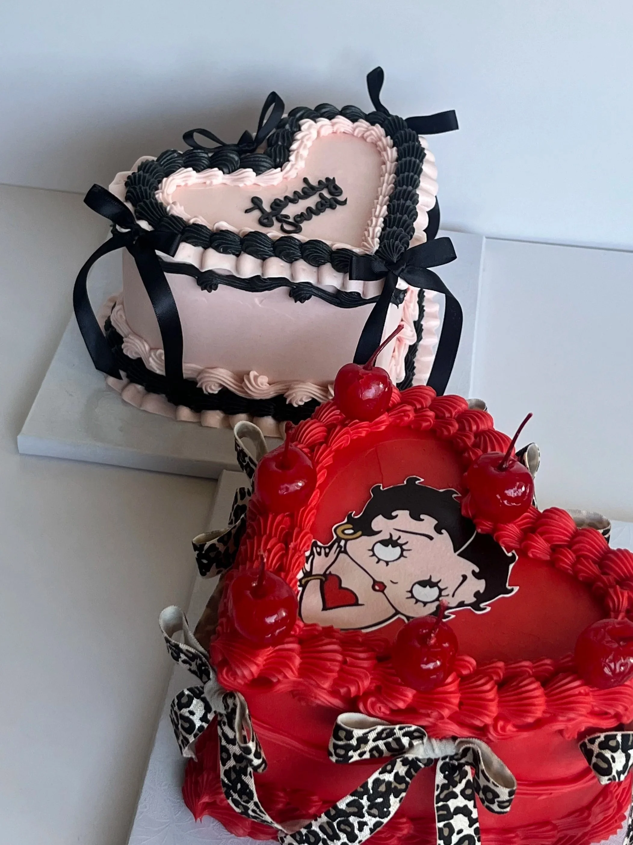 Pink heart-shaped cake with black and pink icing border, black decorative ribbons, and the words 'Jenny James' written on top. There is also a red cake with a Betty Boop character design and red cherries on top.