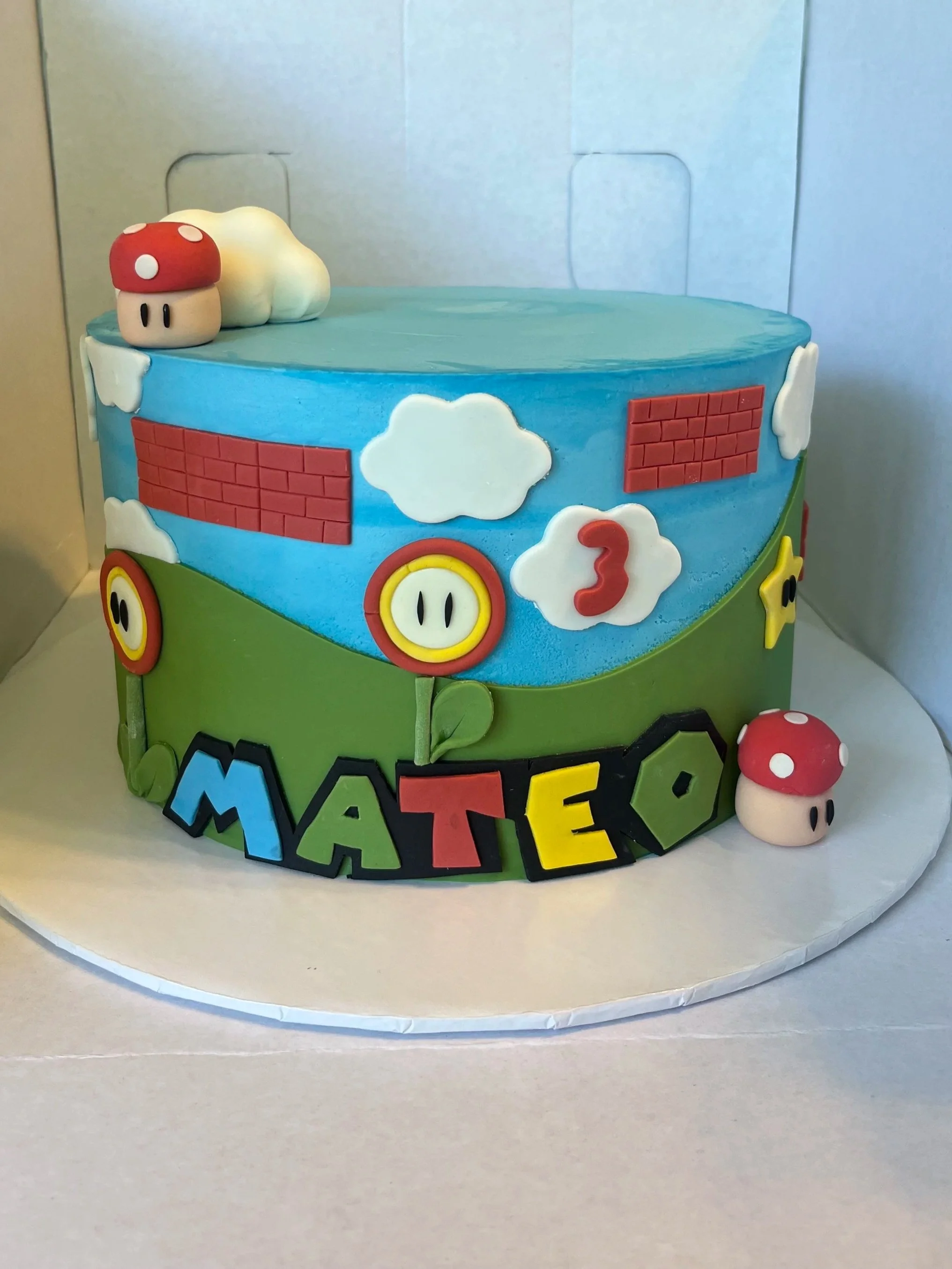 Decorative birthday cake themed on Super Mario, featuring a blue and green color scheme, with Mario's mushroom character on top and colorful 'MATEO' lettering around the base.
