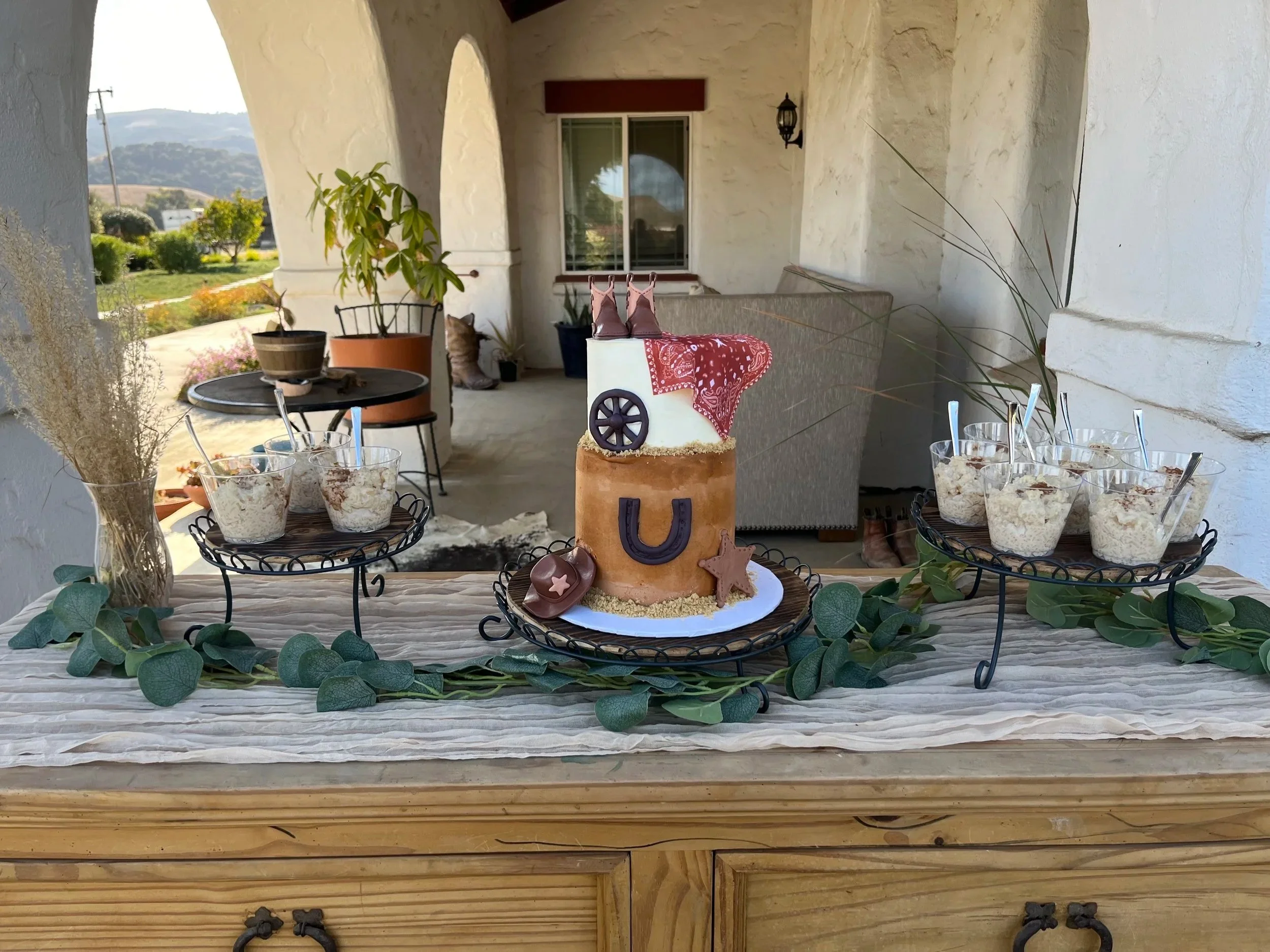 A birthday cake decorated with cowboy themed fondant, pink ballet shoes, and star decorations, placed on a table with glasses of dessert, green leafy garland, and surrounded by chairs and plants on a covered outdoor patio.