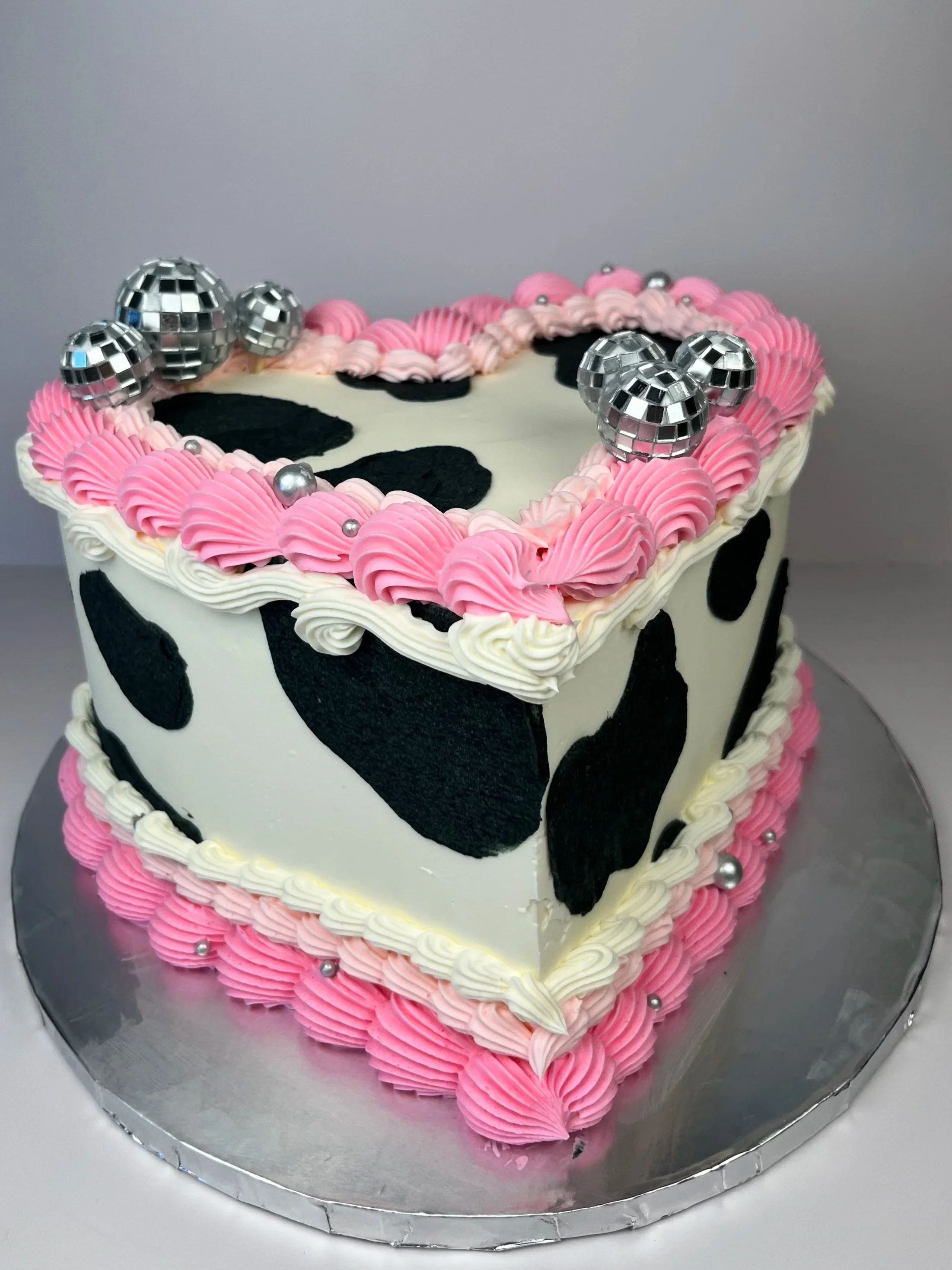 A heart-shaped cake decorated with pink and white frosting, black cow pattern, and silver disco ball cake toppers on a silver cake board.