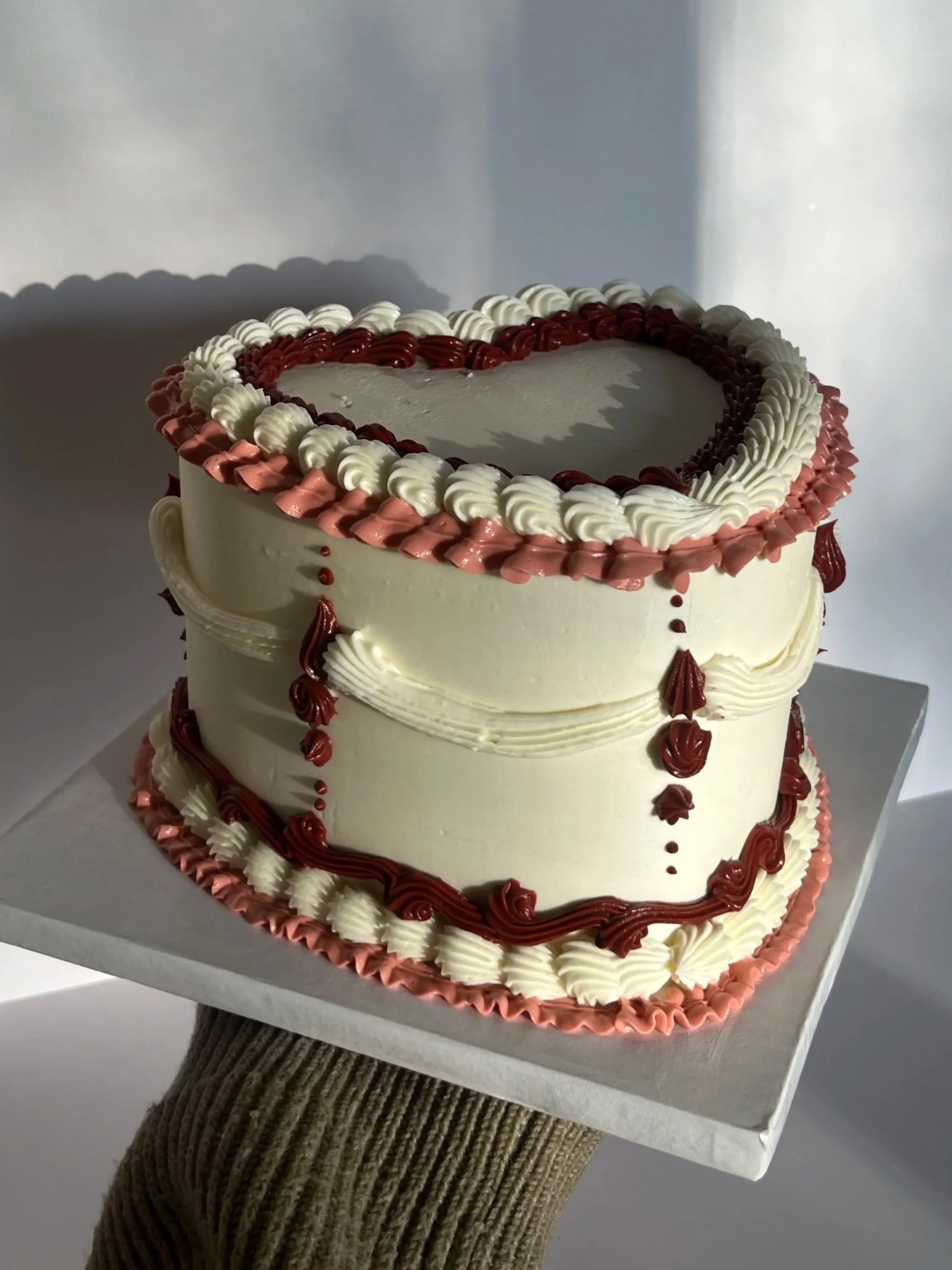 A two-tiered rectangular cake decorated with white, pink, and red frosting with piped borders along the edges and icing drops hanging from the sides, placed on a white cake board.