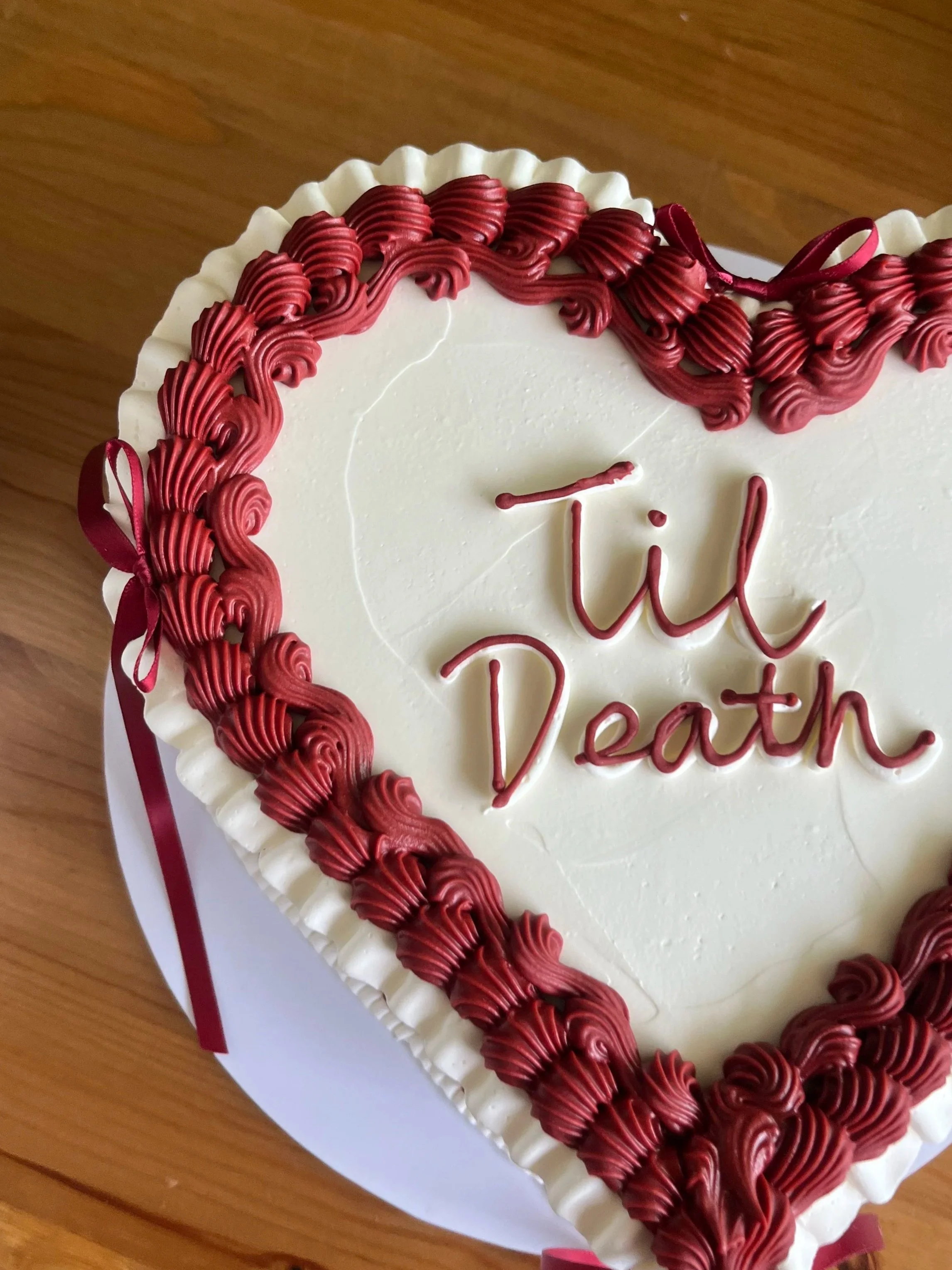 A heart-shaped cake decorated with white icing and red piping along the edges, with a red ribbon on the side and handwritten text on the surface.