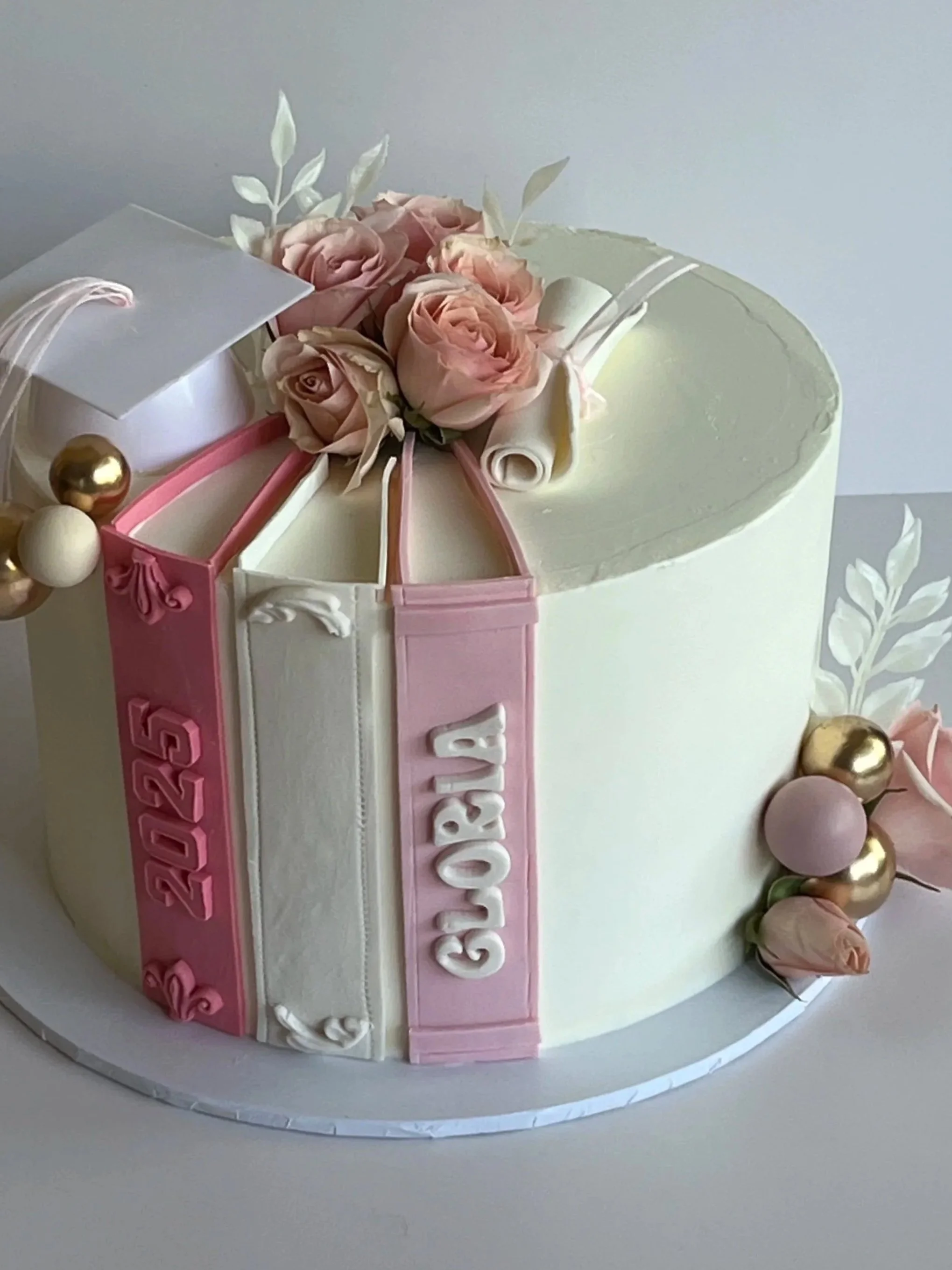 Round white cake decorated with pink and white roses, white leaves, gold and pink balls, and pink and white letter decorations spelling 'Baby Girl'.