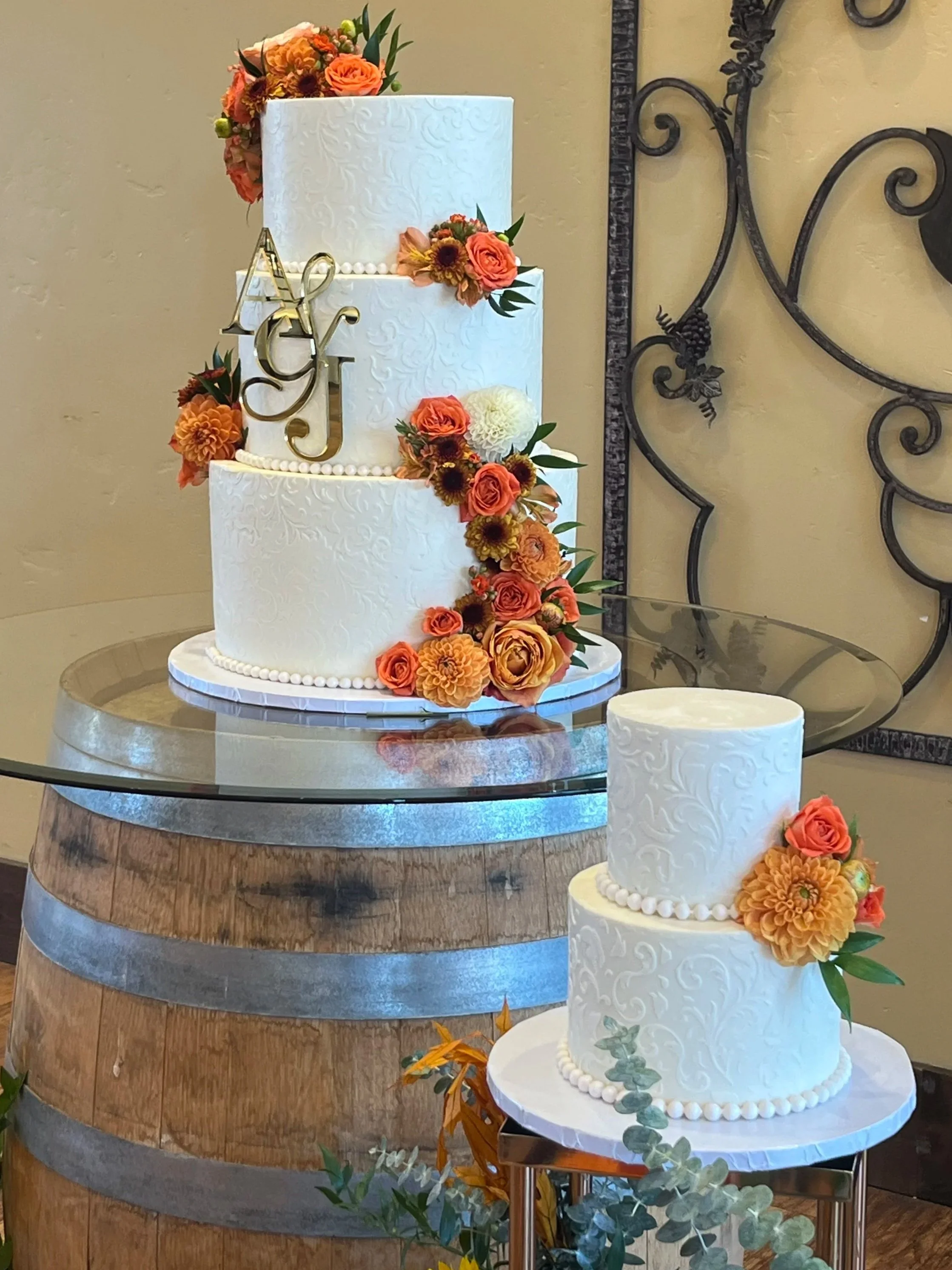Three decorated wedding cakes with floral decorations on a glass table in a room with yellow walls and a large decorative iron wall piece.