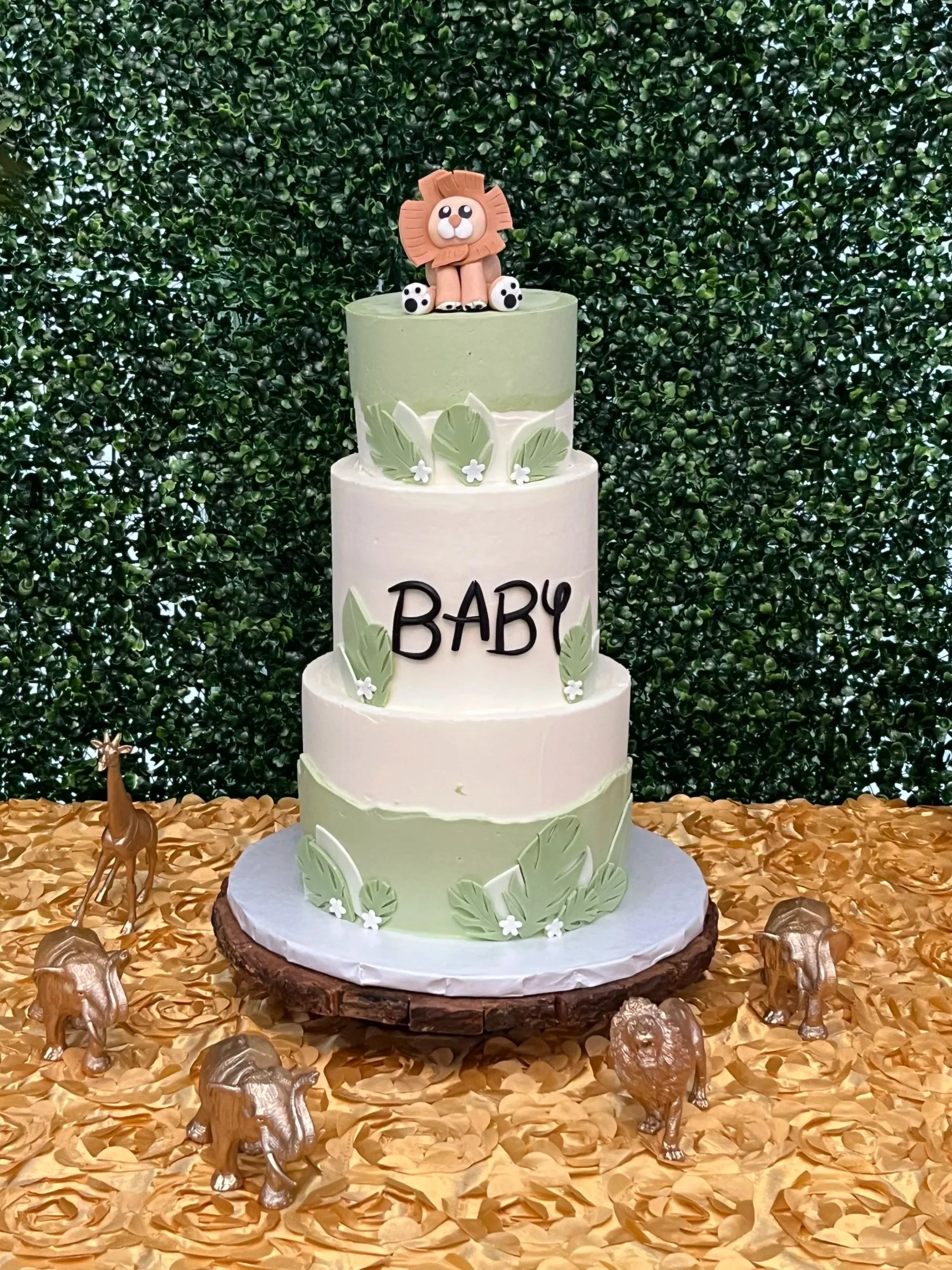 Three-tiered baby shower cake decorated with green leaves and white flowers, topped with a lion figurine, surrounded by small metallic lion figurines on a gold leaf surface with a green leafy background.