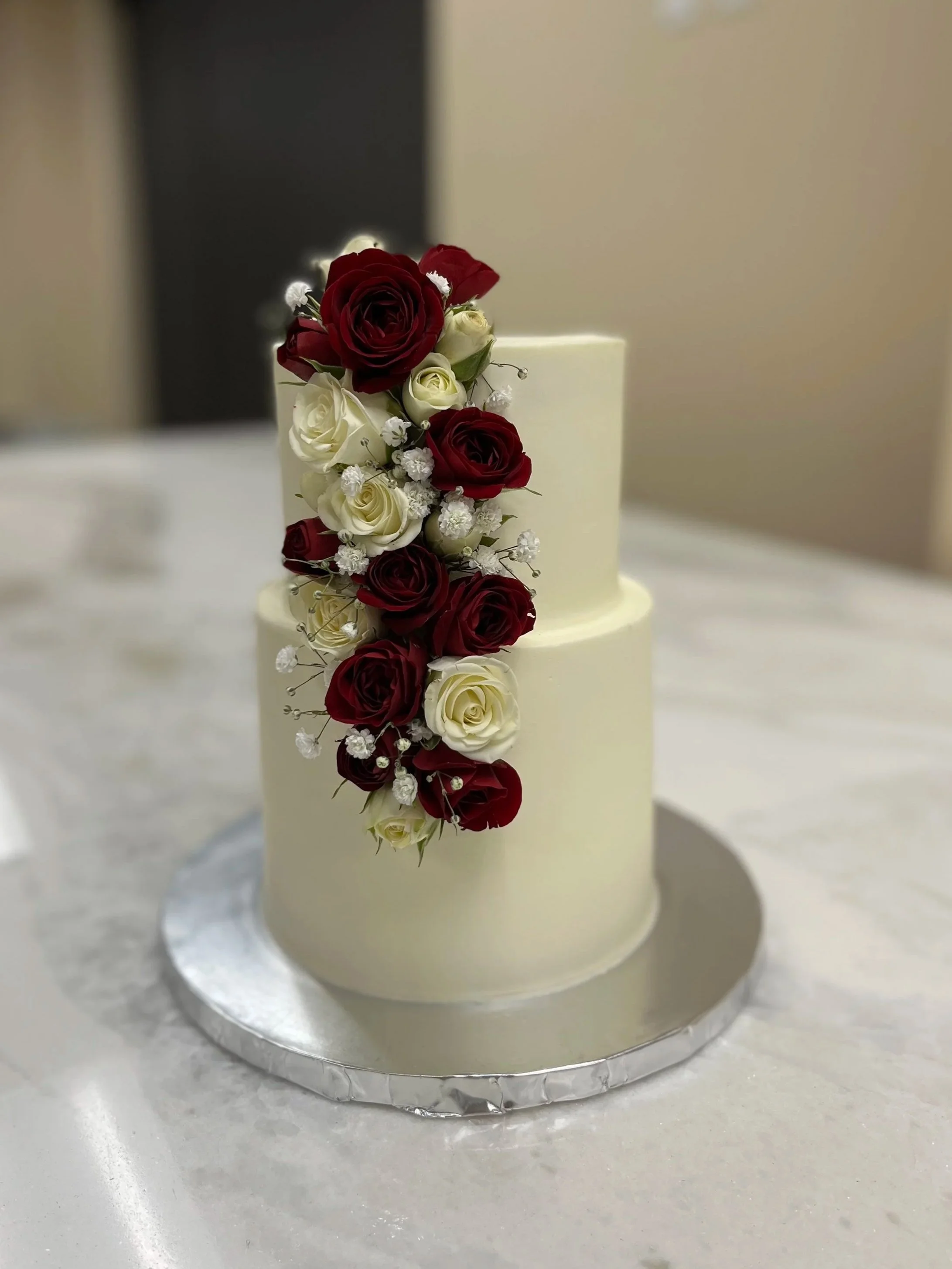 A tall, round white cake decorated with cascading red and white roses and small white flowers on a silver cake board.