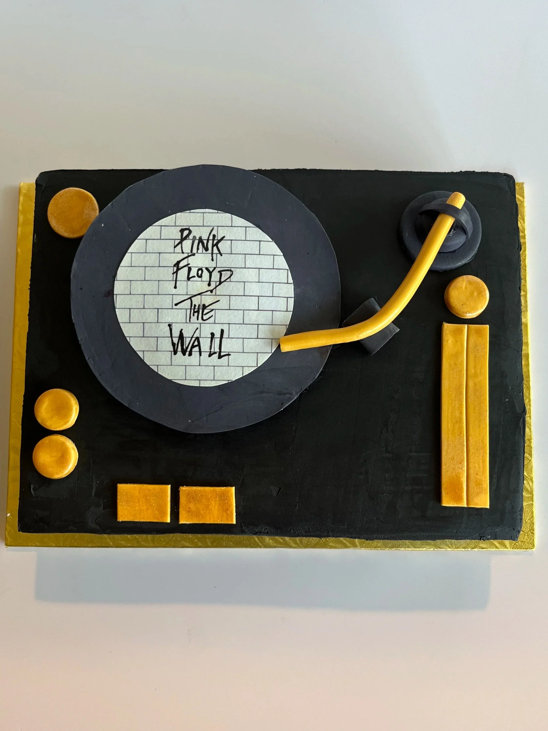A birthday cake decorated in a music theme with a black and gold color scheme. The cake has a circular black decoration with a white background that has the words 'Pink Floyd The Wall' written on it. It is topped with a yellow guitar made of cake or 