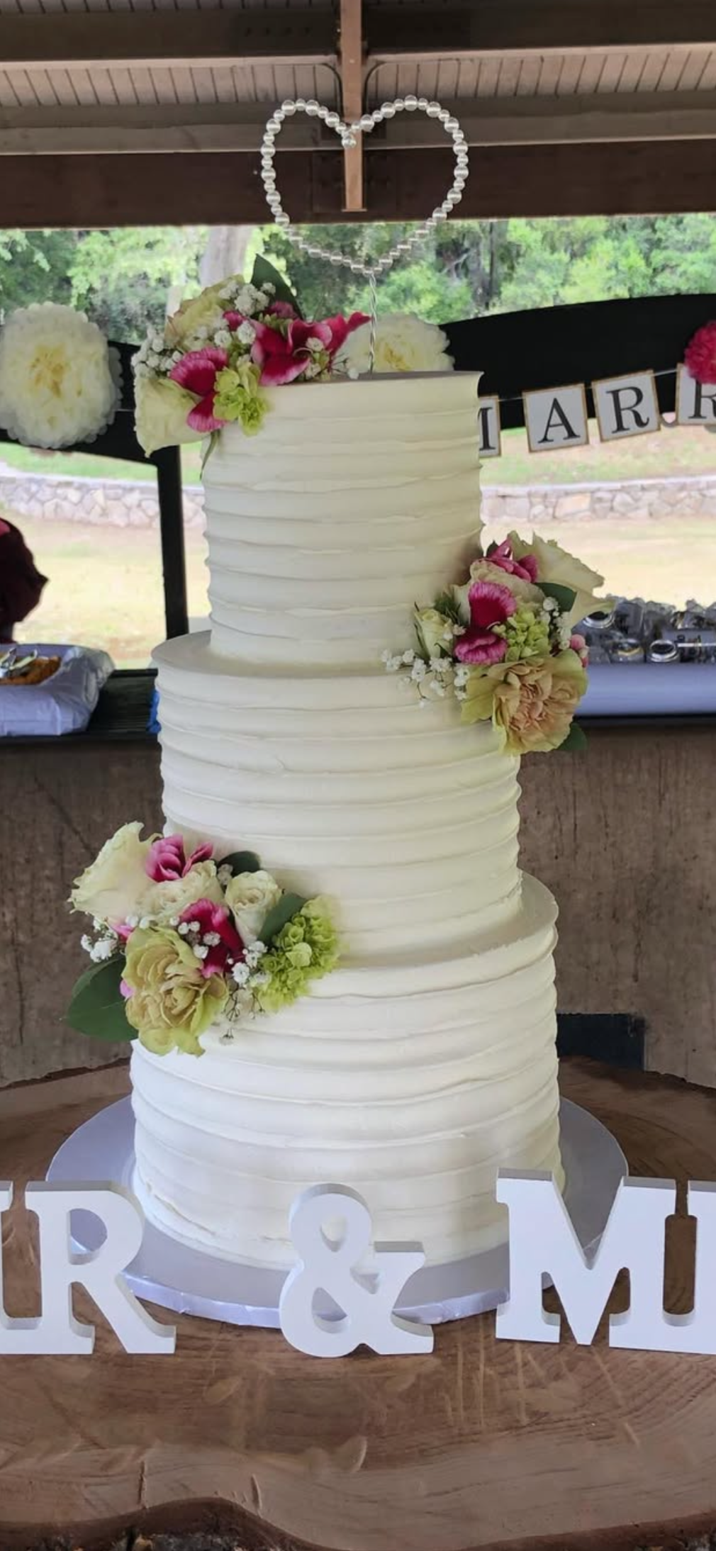 A multi-tiered white wedding cake decorated with pink, white, and green flowers, topped with a heart-shaped bead decoration, on a wooden table with 'MR & MRS' white letters in front.