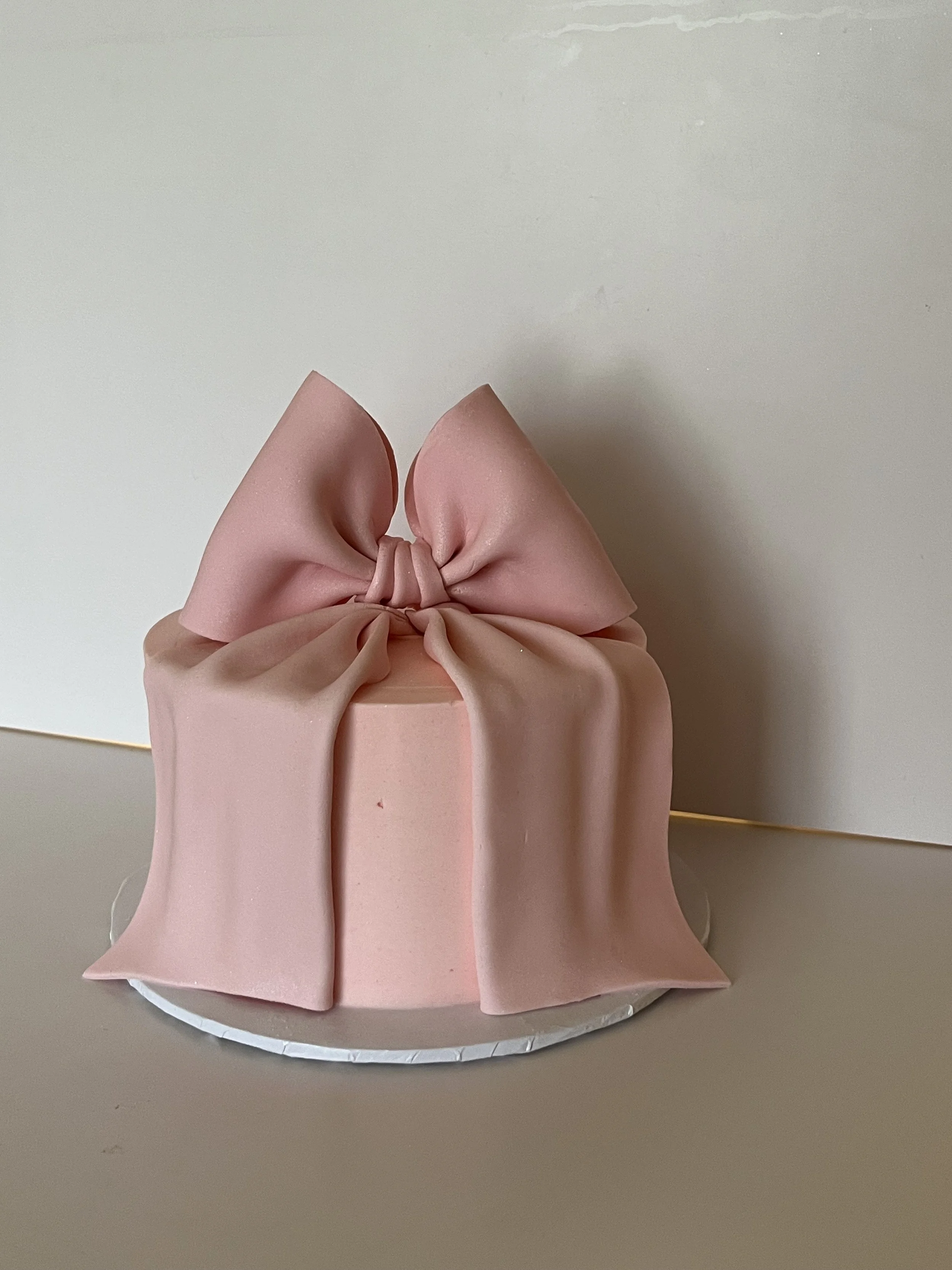 Pink cake with a bow-shaped decoration on top, placed on a white round platter against a plain wall background.